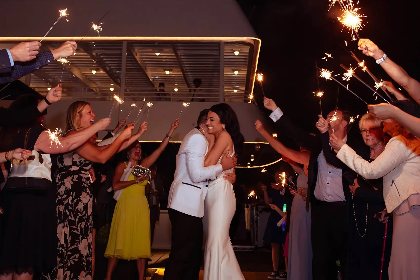 A bride and groom dancing and hugging in the center of a celebration as guests hold sparklers around them at night.