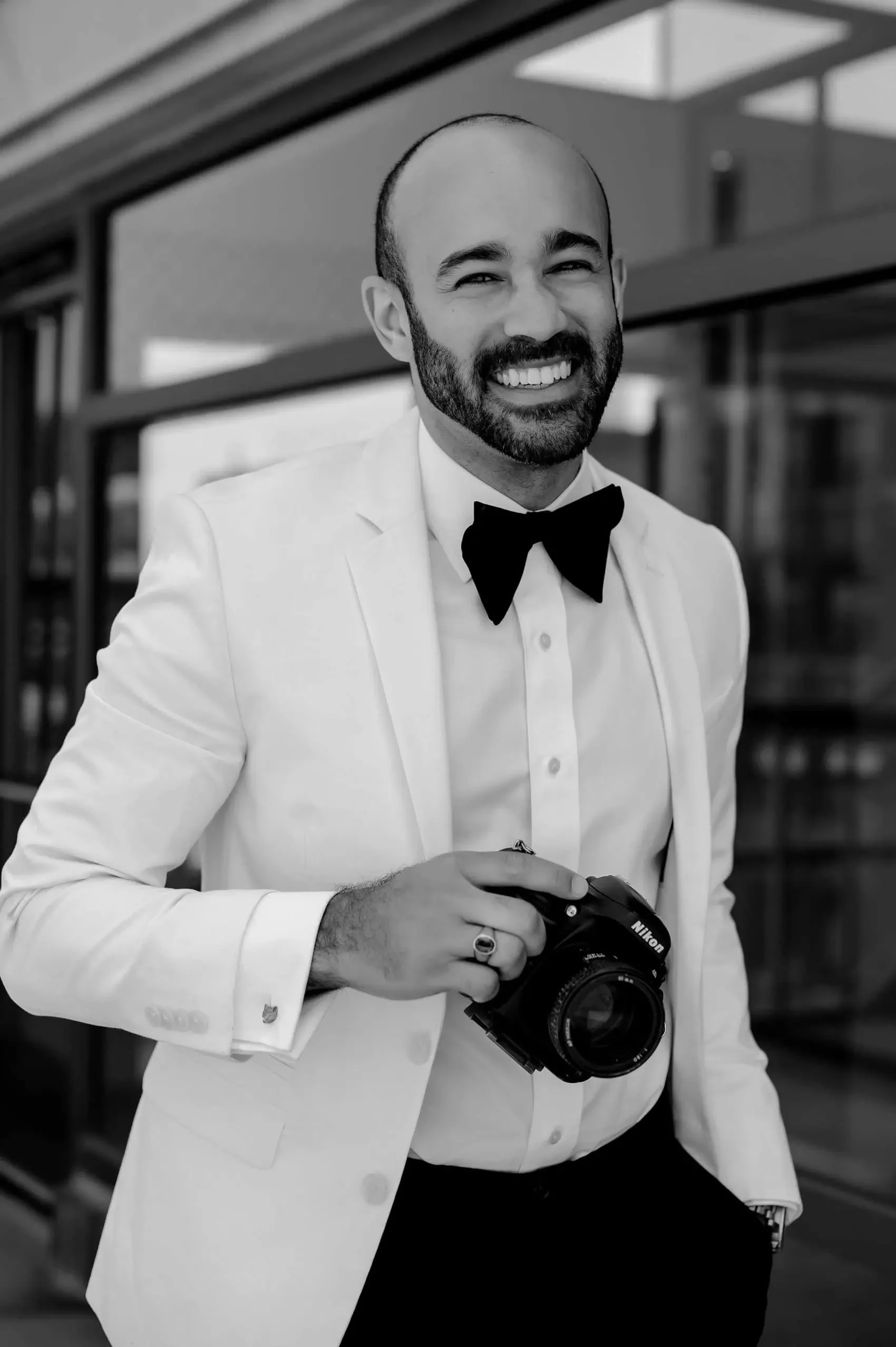 A man dressed in a tuxedo with a bow tie, smiling and holding a Nikon camera in front of a modern building.