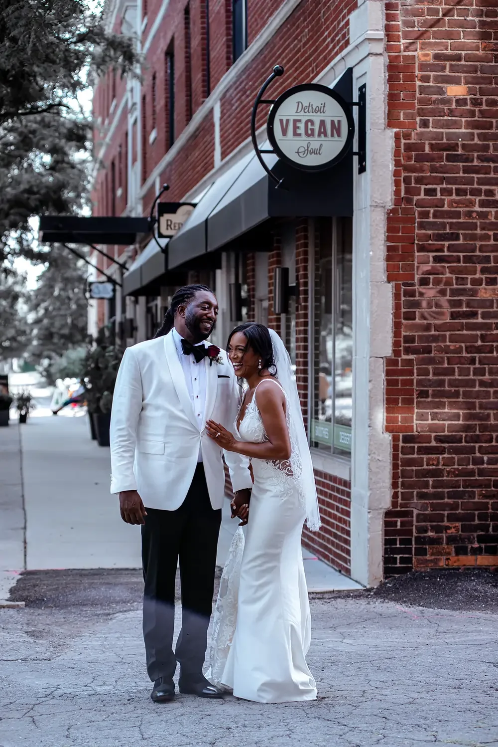 A bride and groom on a city sidewalk, smiling and holding hands outside a restaurant with a sign that reads "Detroit Vegan Soul."