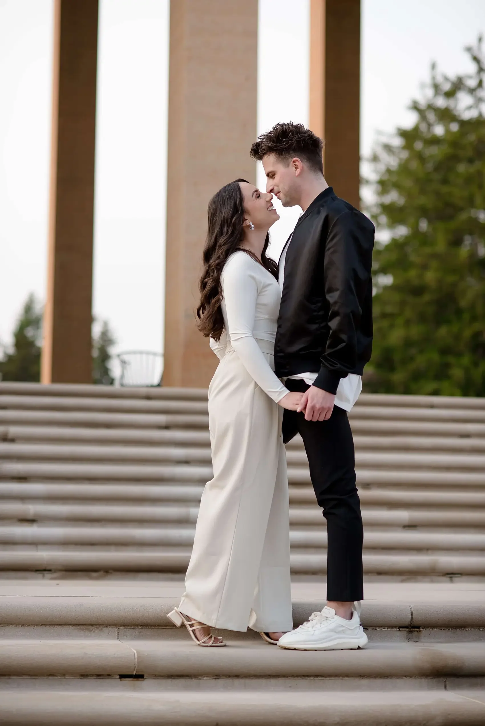 A young couple standing close, holding hands and leaning their foreheads together, smiling, on the steps in front of a large architectural structure with trees in the background.
