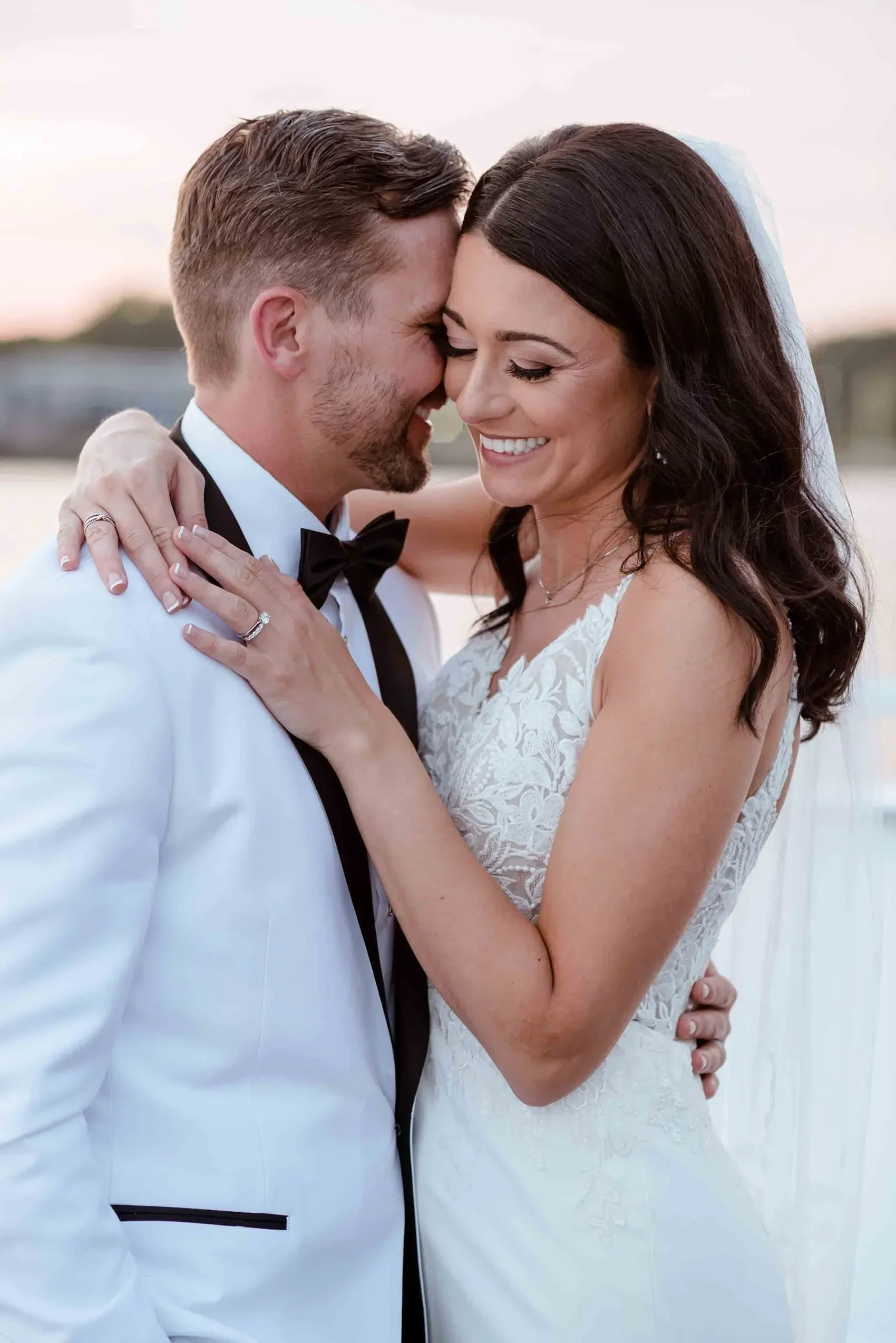 A newlywed couple embracing, smiling with foreheads pressed together outdoors during sunset.