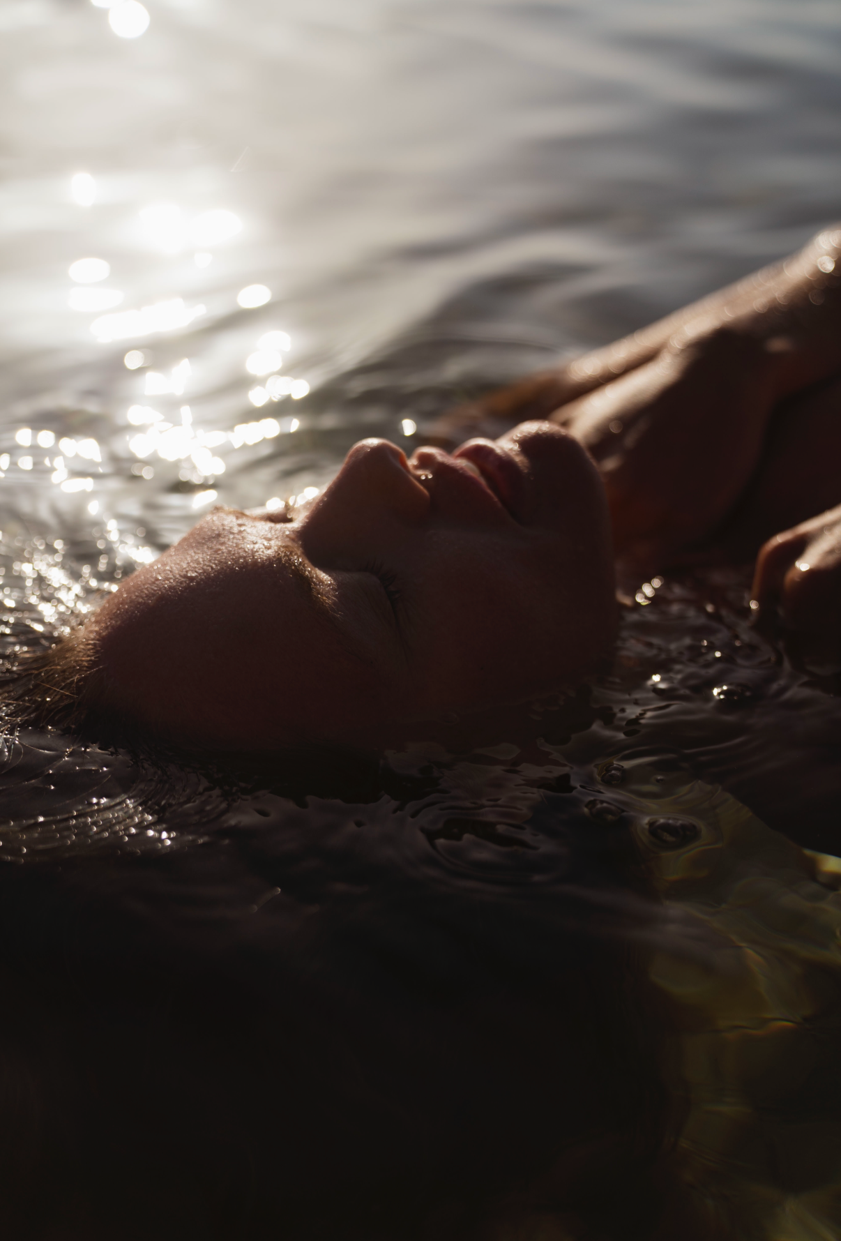 Person lying in water with face partially submerged, sunlight reflecting on water surface.