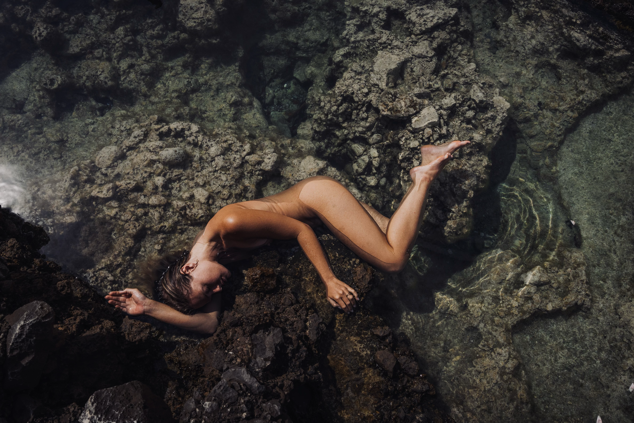 A naked woman lying on her side on a rocky shoreline with water around her.