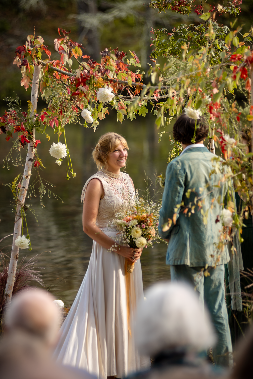 A woman in a white dress holding a bouquet of flowers, smiling at a man in a light-colored suit during a wedding ceremony outdoors by a body of water.