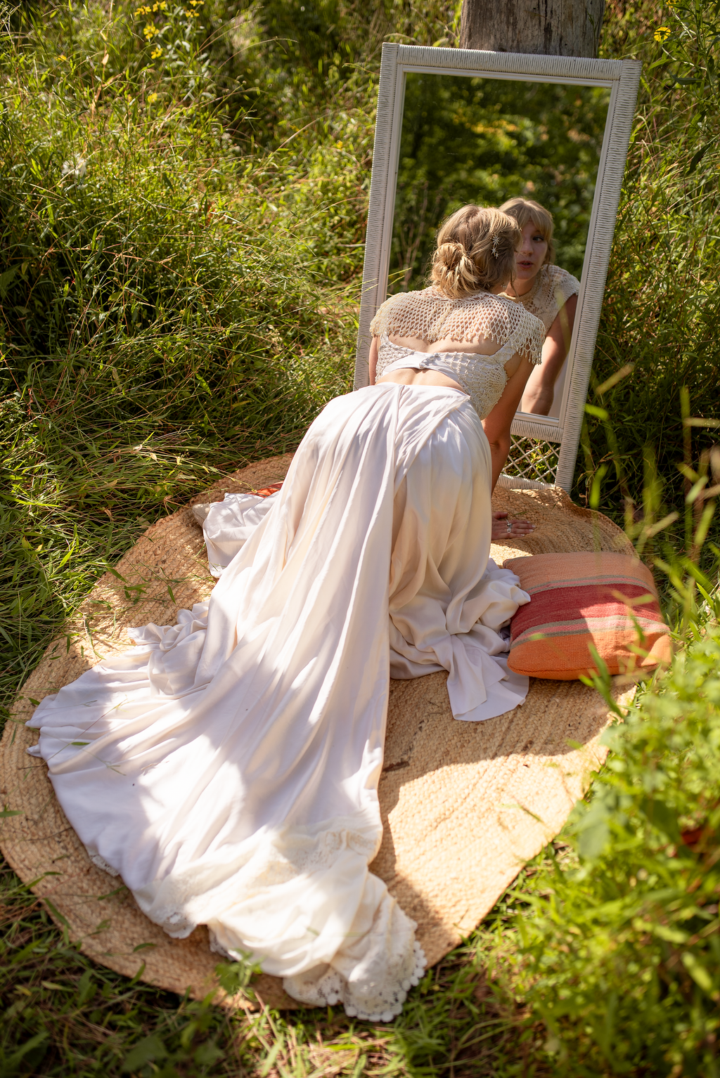 A woman in a vintage white dress and lace top is kneeling on a woven rug with a pillow, looking into a mirror outdoors among tall grass and trees.