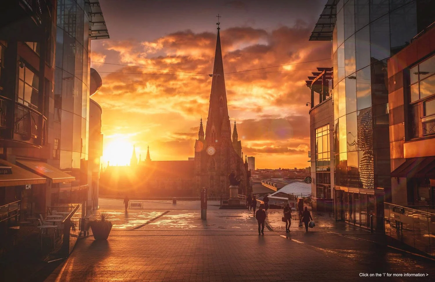 Sunset or sunrise over a city square with a historic church or cathedral in the background, framed by modern glass buildings on both sides, with a few people walking and some reflections on the glass surfaces.