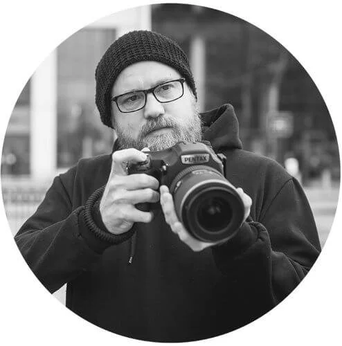 Black and white photo of a man wearing glasses, a beanie, and a hoodie, holding a DSLR camera and looking at the viewer