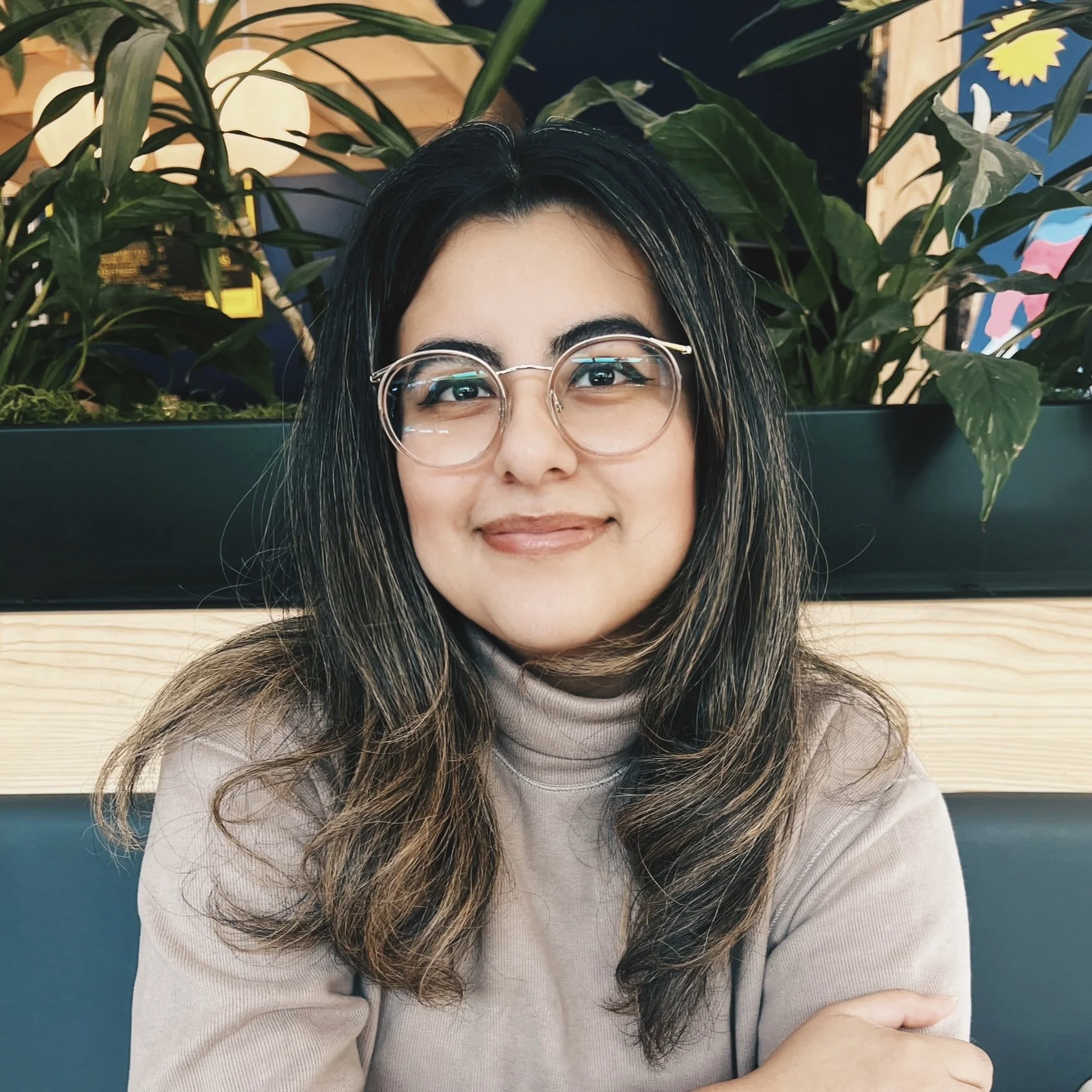A smiling woman with long dark hair and glasses sitting in front of green plants in a modern indoor setting.