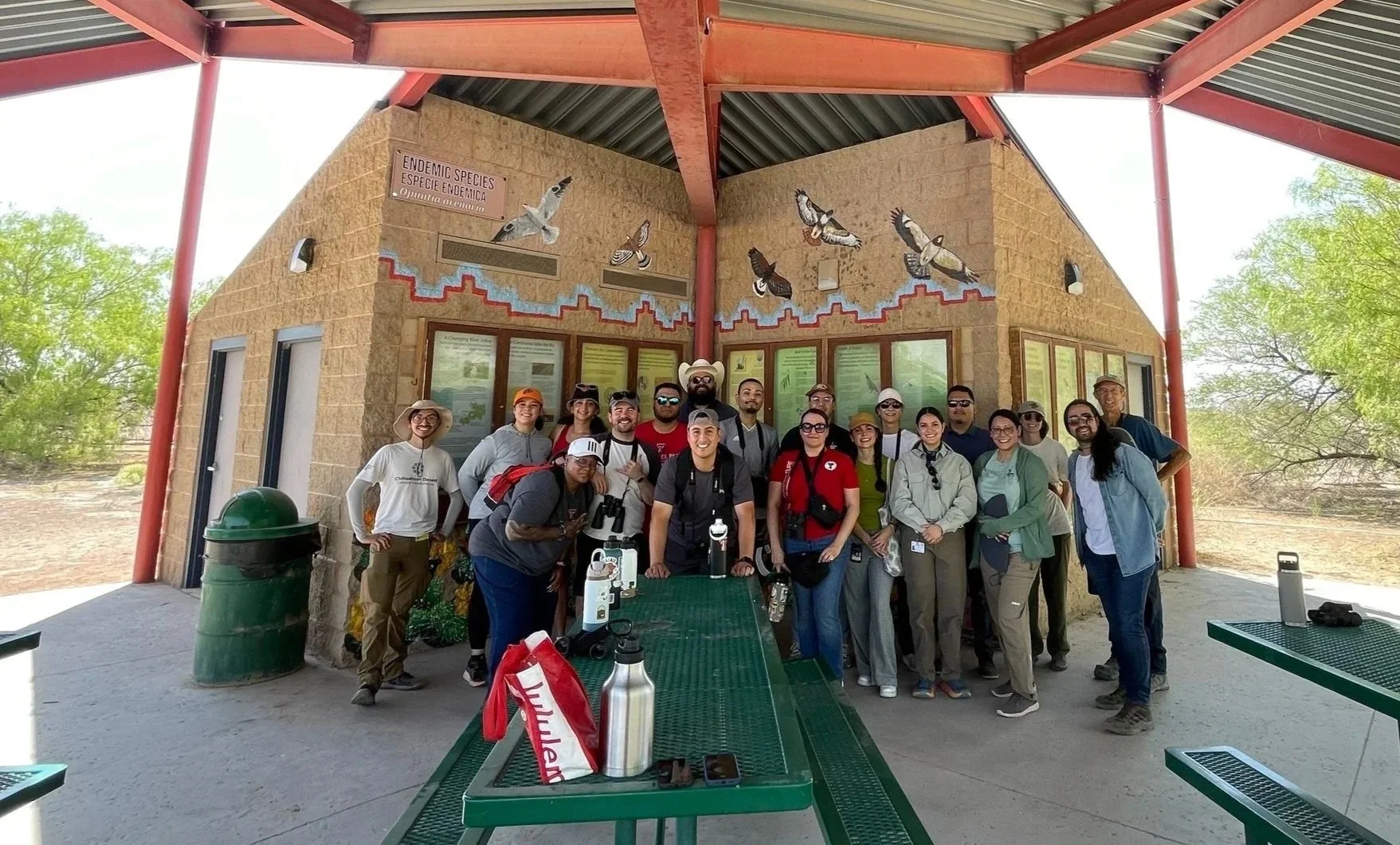 Group of people standing together outdoors in front of an educational building with bird illustrations, under a metal roofed shelter, with some trees and desert landscape in the background.