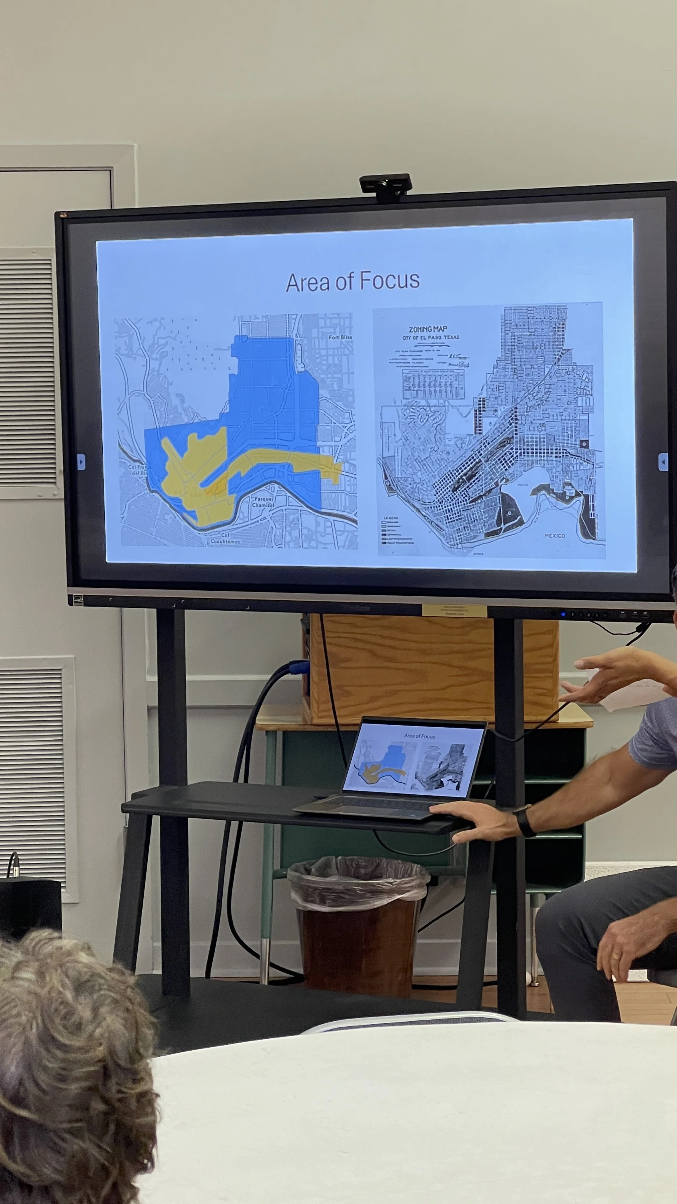 Presentation screen showing maps titled 'Area of Focus,' with one map highlighting regions in blue and yellow, and another map of an urban area. A person is pointing at the screen, and a laptop connected to the display is on a stand.