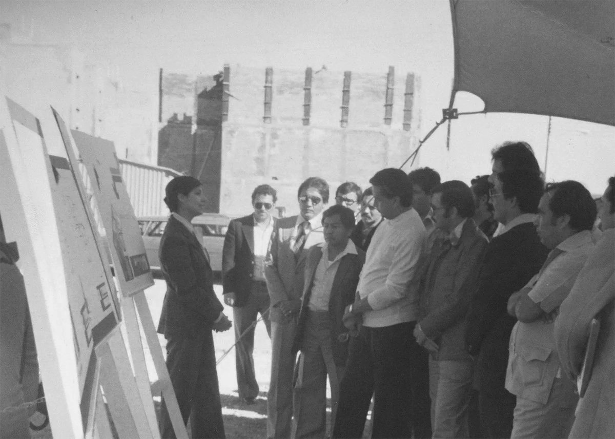 Black and white photo of a woman giving a presentation to a group of men outdoors, with posters or boards displayed nearby.
