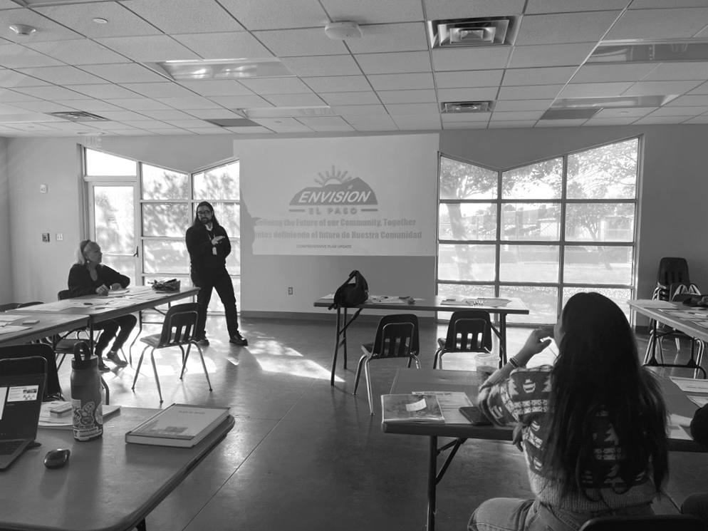 A classroom with students and a teacher, some students sitting at desks and one woman standing near a projector screen, in a room with large windows and ceiling lights.