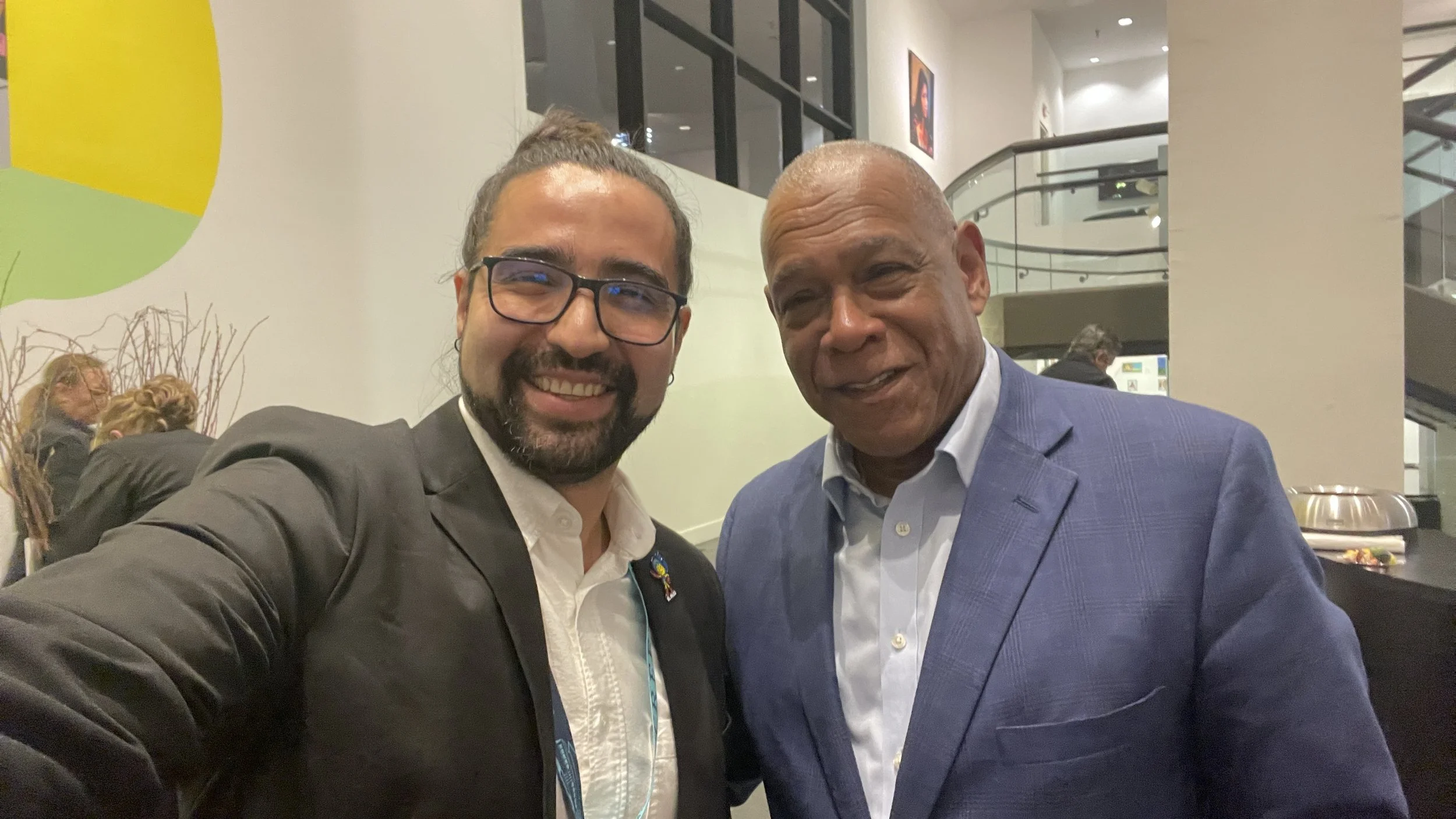 Two men smiling and taking a selfie together in an indoor setting. The man on the left has glasses, a beard, and is wearing a dark blazer and white shirt. The man on the right is bald and wearing a blue blazer with a light-colored shirt.