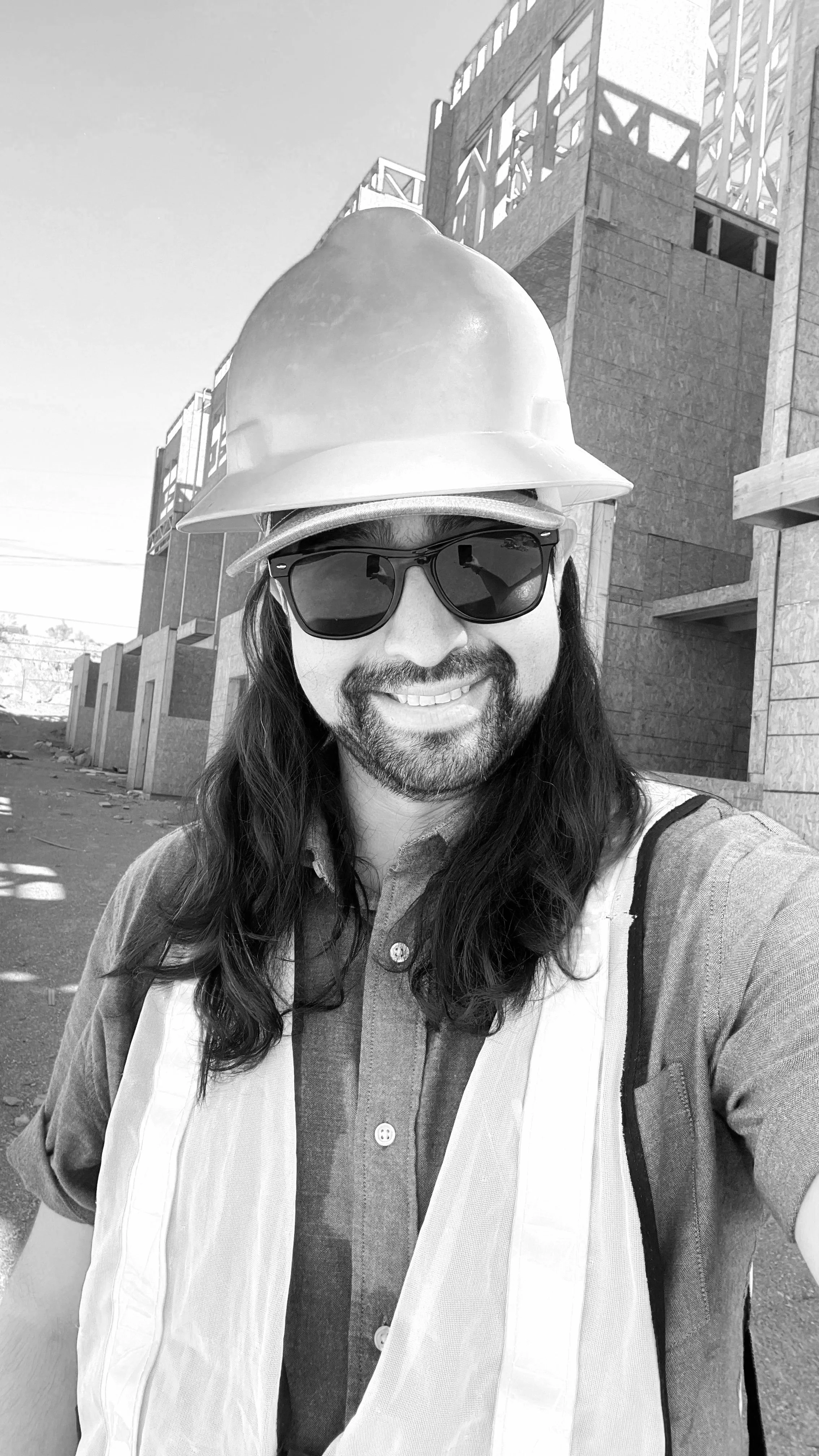 A man with long hair and a beard wearing sunglasses, a hard hat, and a safety vest at a construction site.