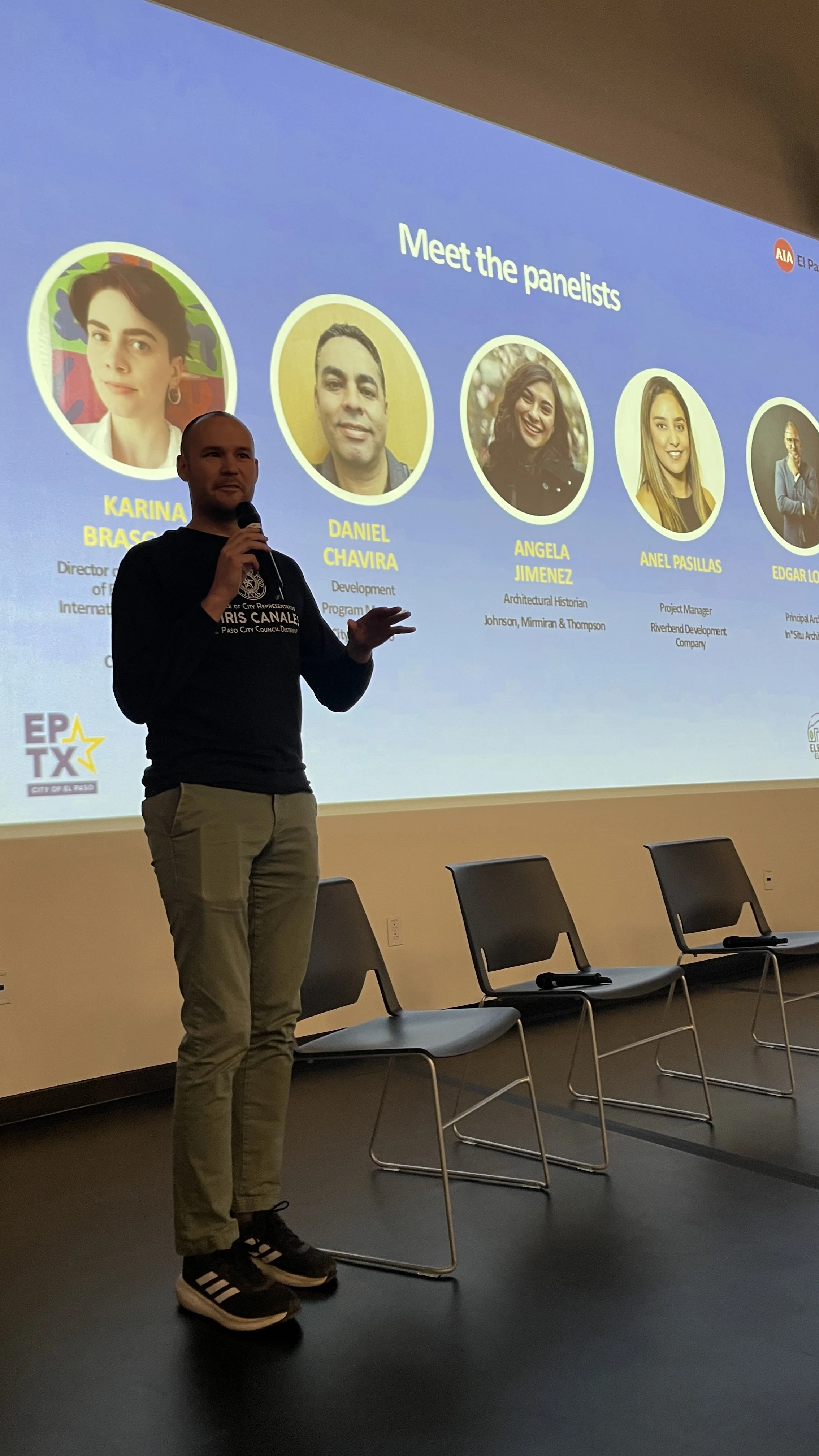A man standing on a stage with a microphone in front of a large screen displaying panelists' photos and names during a presentation or panel discussion.