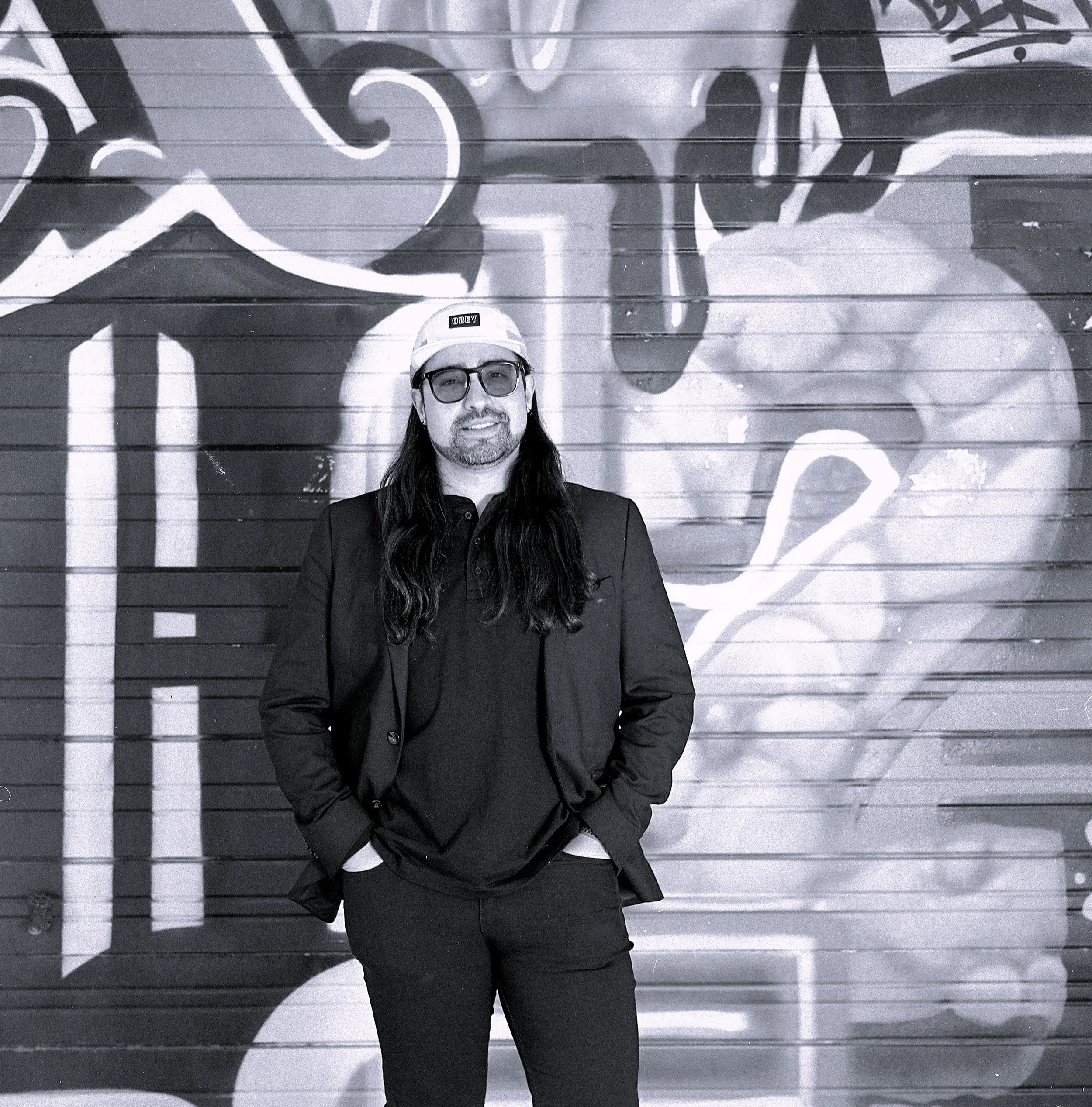 Man with long hair, beard, sunglasses, black jacket, and cap standing in front of graffiti wall.