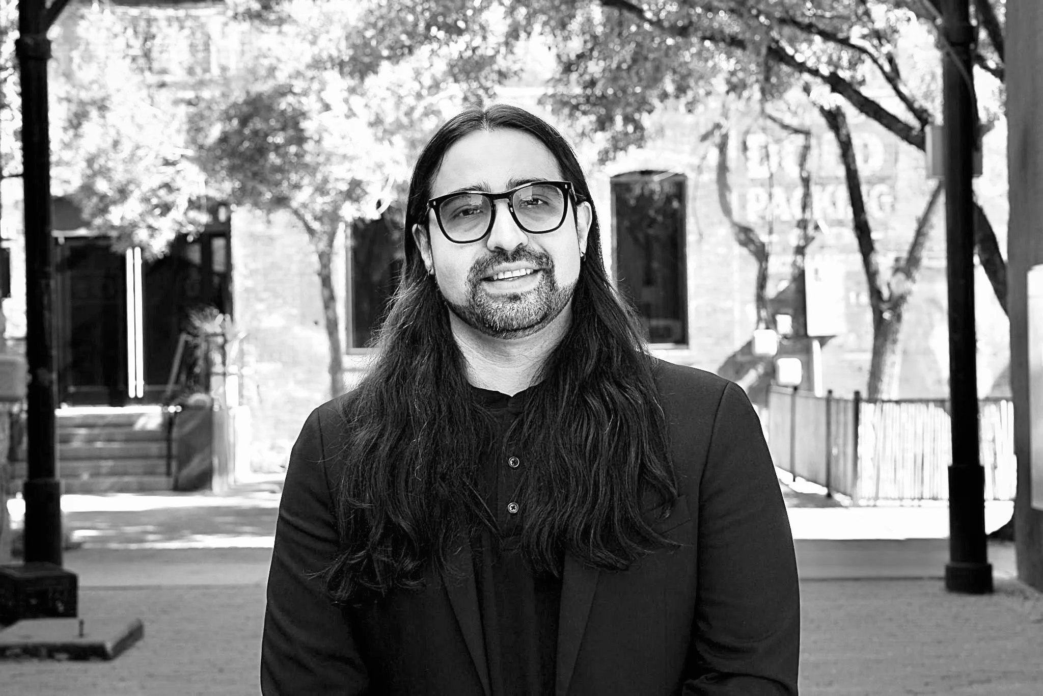 Black and white photo of a man with long dark hair, glasses, a beard, wearing a dark blazer, standing outdoors with trees and buildings in the background.