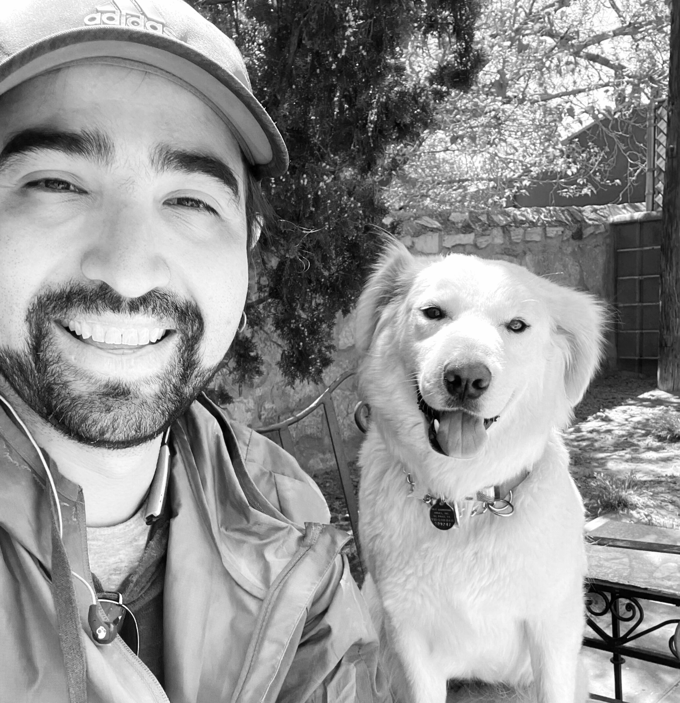 A man smiling and a happy dog sitting outdoors, with trees and a stone wall in the background.