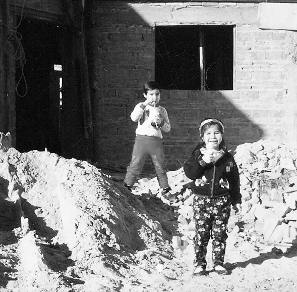 Two children, a boy and a girl, standing on a pile of dirt and rubble outside a brick building with a window.