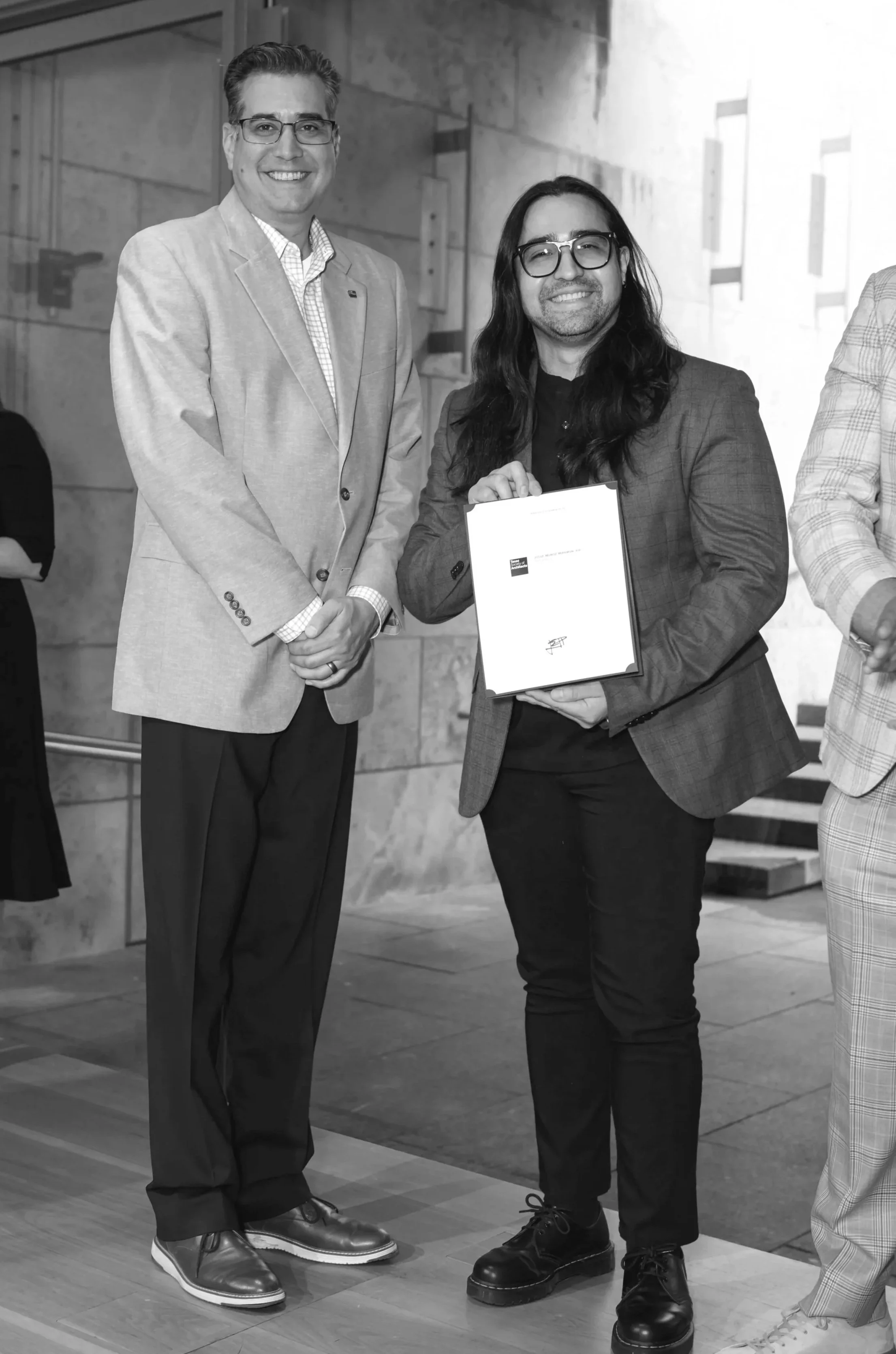 Two men in business attire standing indoors, one of them holding a framed certificate, smiling at the camera.