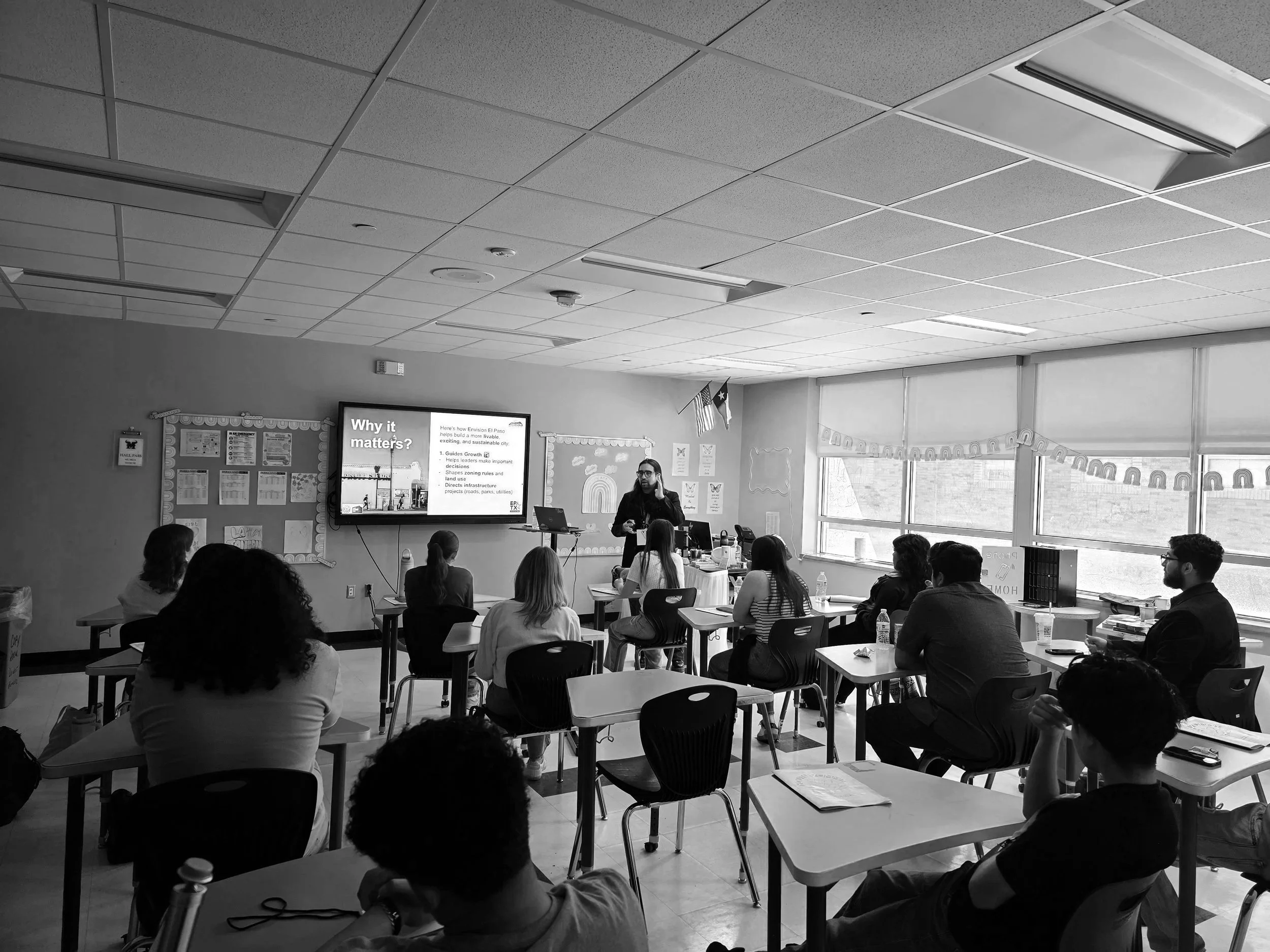 A classroom with students seated at individual desks and a teacher presenting at the front using a large screen. The classroom has large windows on the right side and educational decorations on the walls.