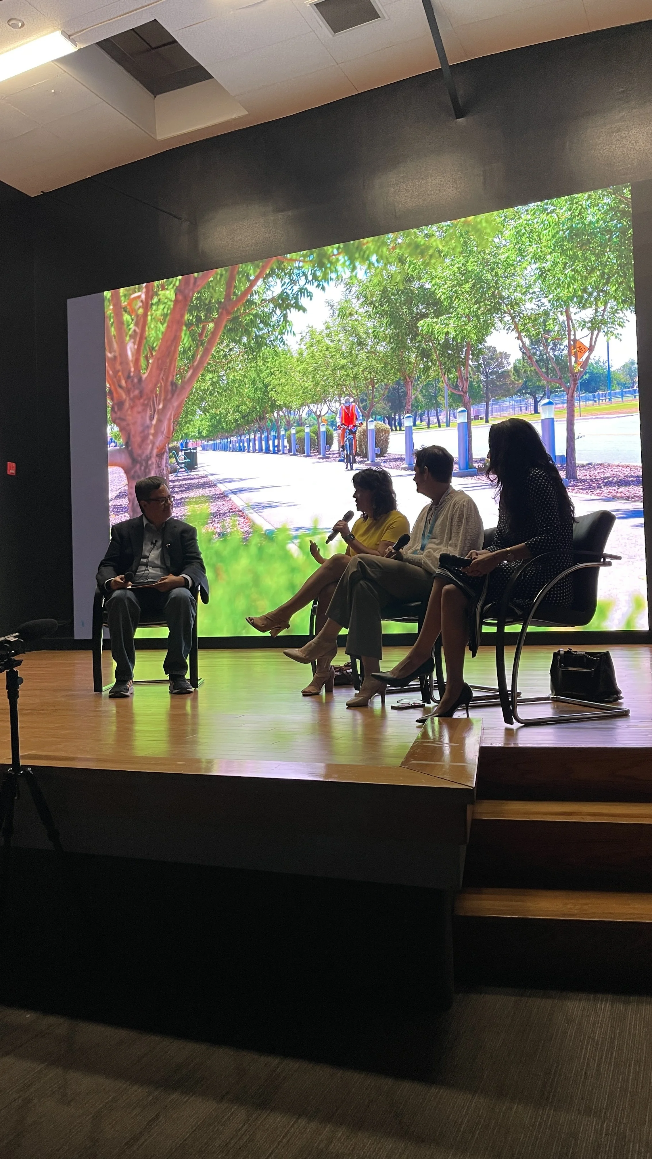 A panel discussion taking place on a stage with four people, three women and one man, seated in chairs. The woman second from the left is speaking into a microphone. Behind them is a large screen showing a scenic outdoor scene with trees, a sidewalk, and a person riding a bicycle.