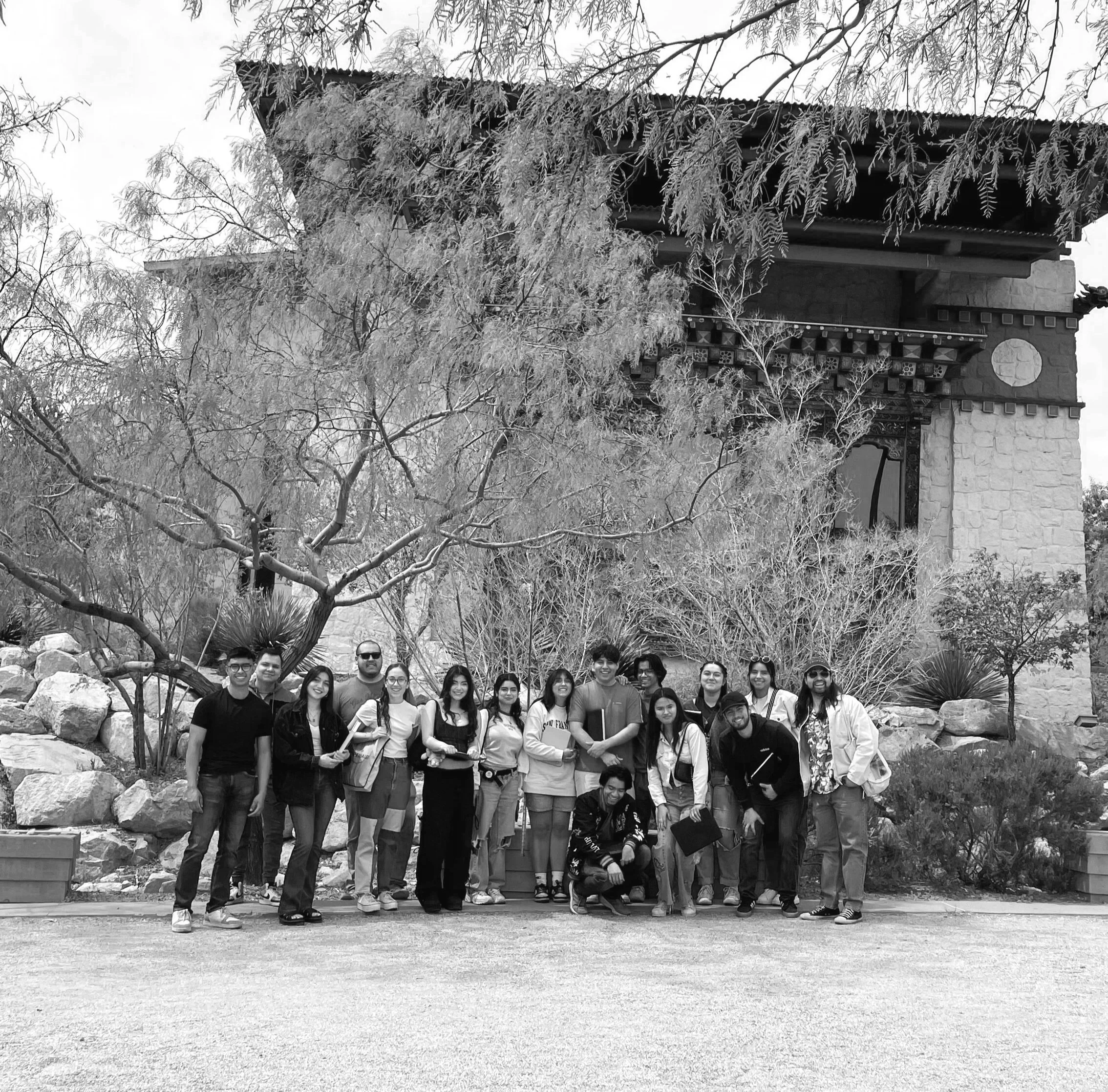Group of people standing outdoors in front of a building with trees and rocks, posing for a photo.