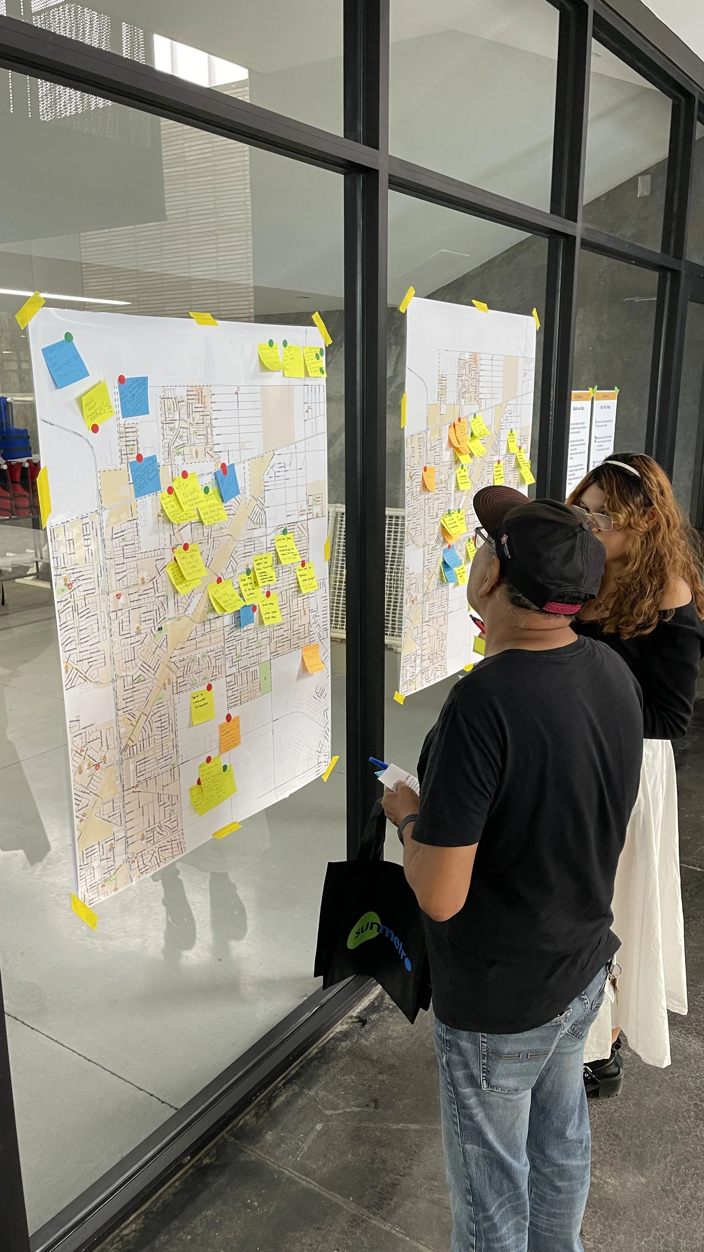 Two people looking at maps on a glass wall, covered with colorful sticky notes and markers in a meeting or workshop setting.