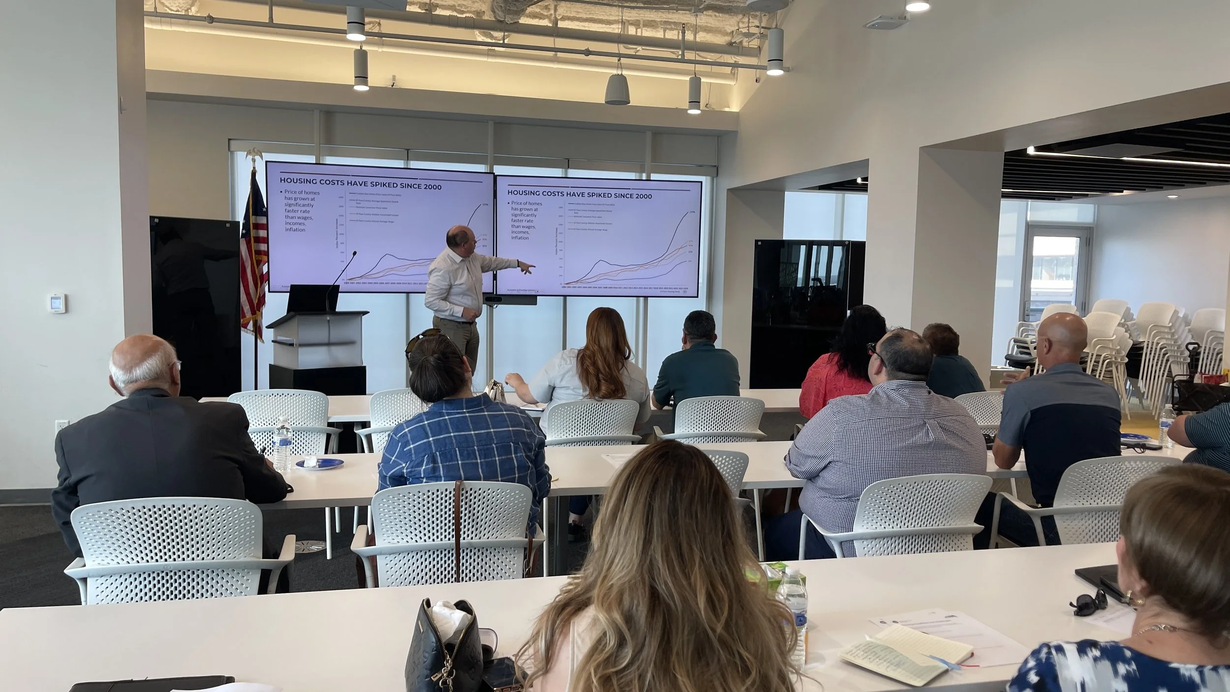 A man in a white shirt is giving a presentation in front of a large screen displaying graphs about housing costs. The audience is seated at white tables, attentively listening. The room has modern lighting and a window in the background.