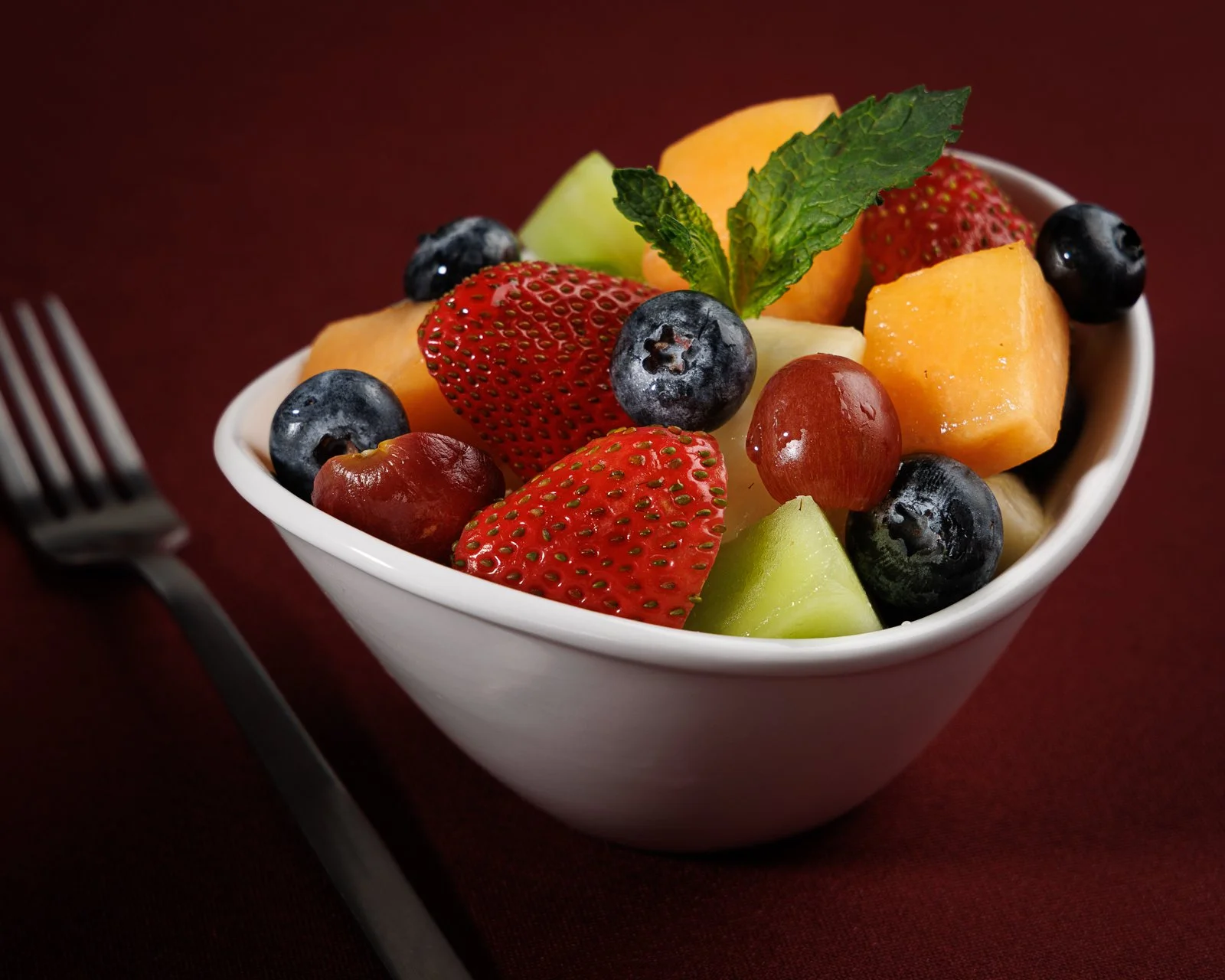 A white bowl filled with mixed fresh fruit including strawberries, blueberries, cantaloupe, honeydew, grapes, and a sprig of mint, placed on a dark red surface with a fork beside it.