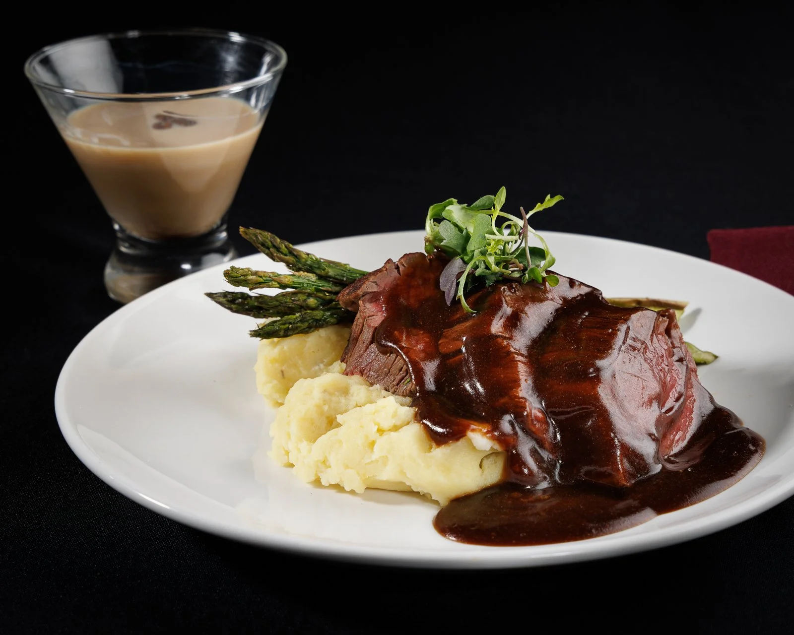 Plate of mashed potatoes with asparagus and a beef steak topped with brown gravy and microgreens, served alongside a glass of creamy brown sauce on a black background.