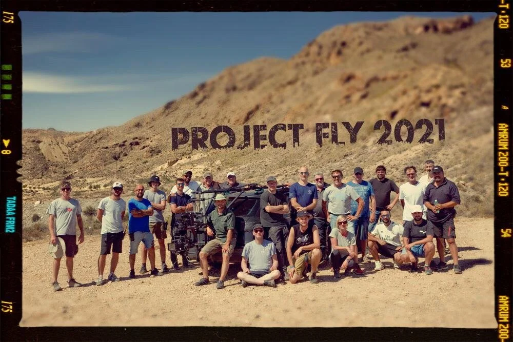 Group of people posing outdoors in a desert landscape with mountains, with text reading 'PROJECT FLY 2021' in the background.