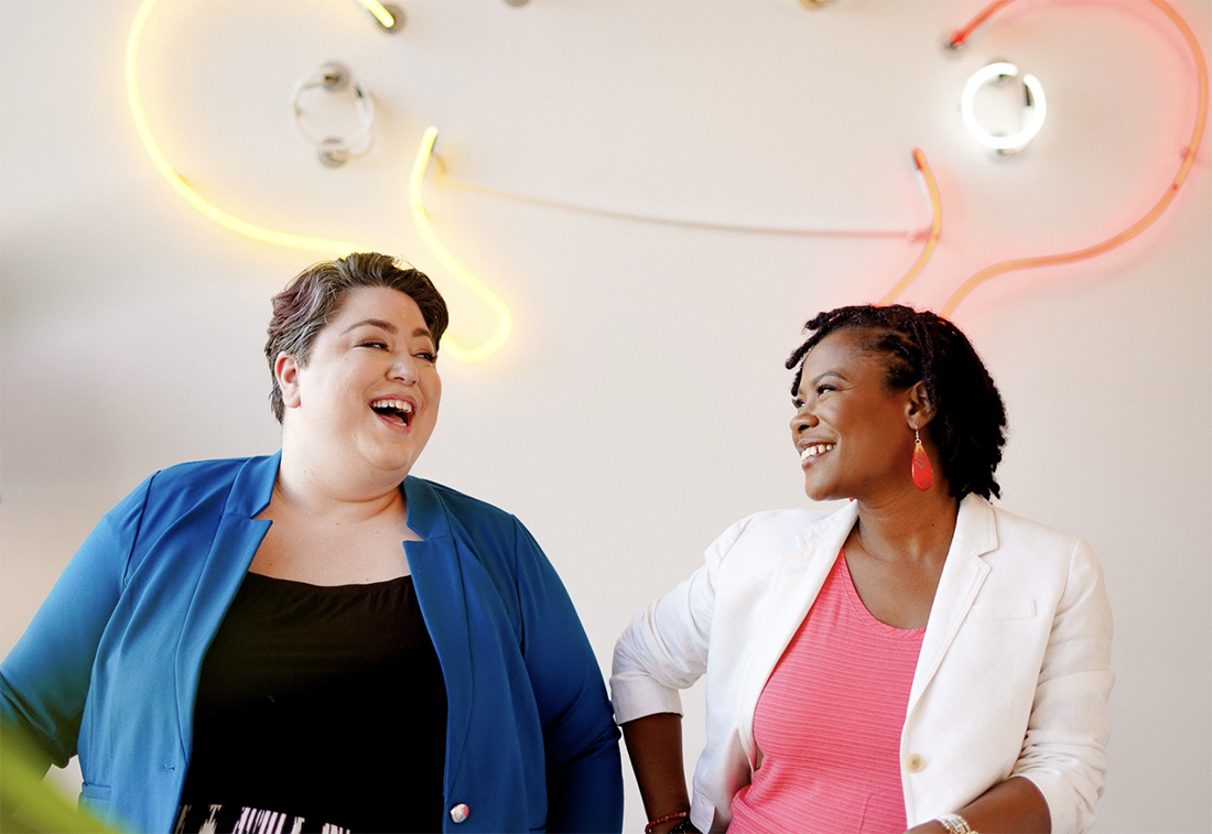 Two people laughing in front of neon art on a wall.