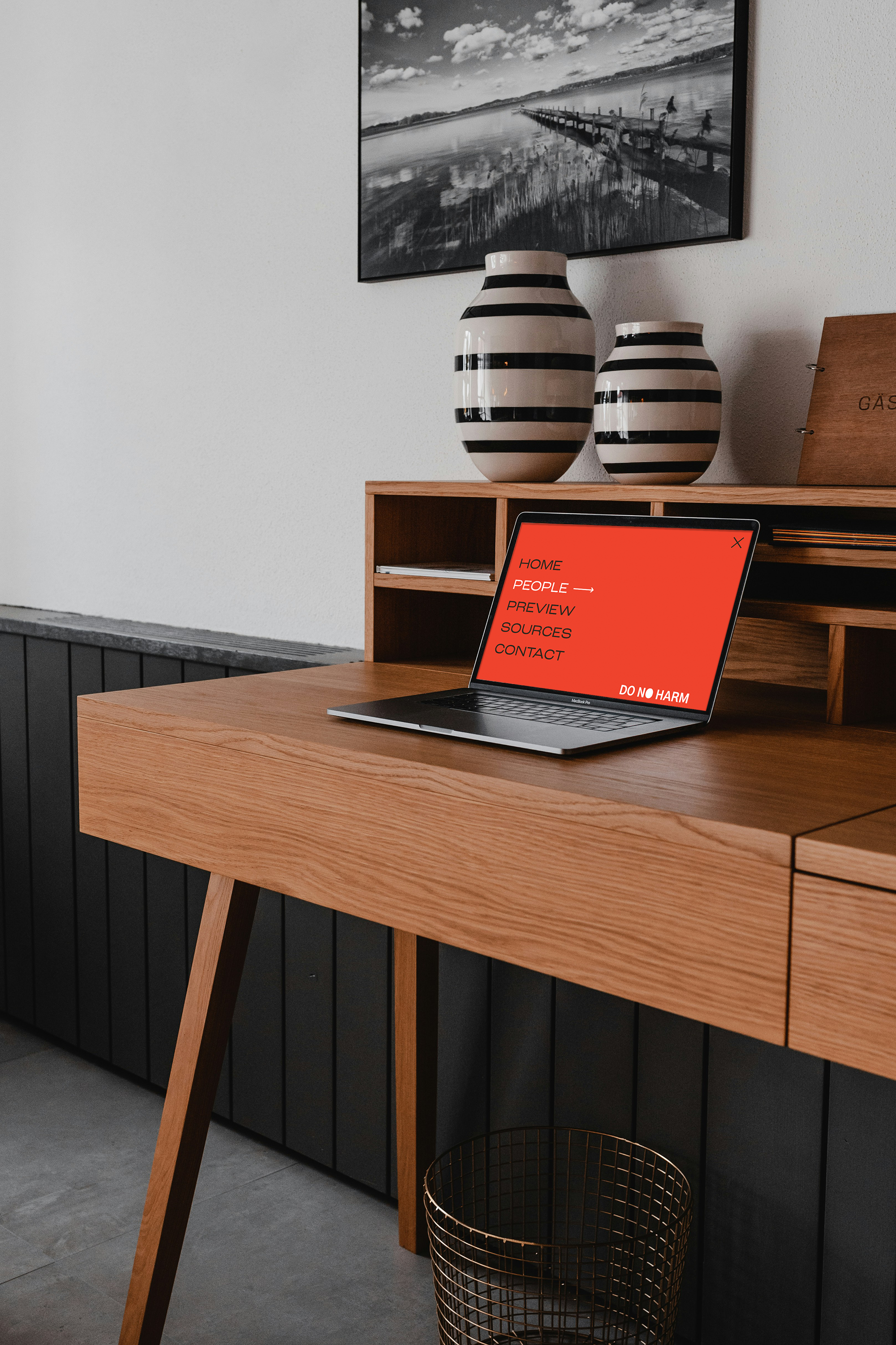 Wooden desk with a laptop displaying a red screen, two striped vases, a black and white photograph above, and a metal wastebasket below.