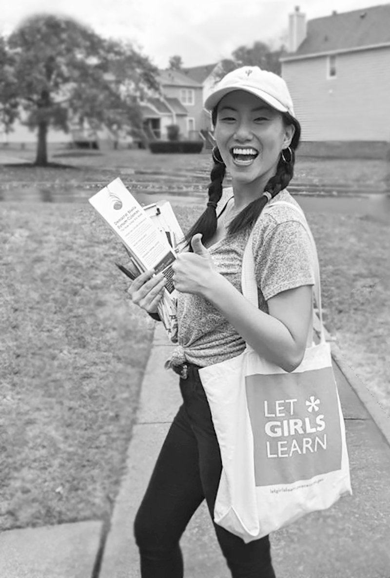 Person smiling and holding a "Let Girls Learn" tote bag, giving a thumbs-up, standing outdoors on a sidewalk.