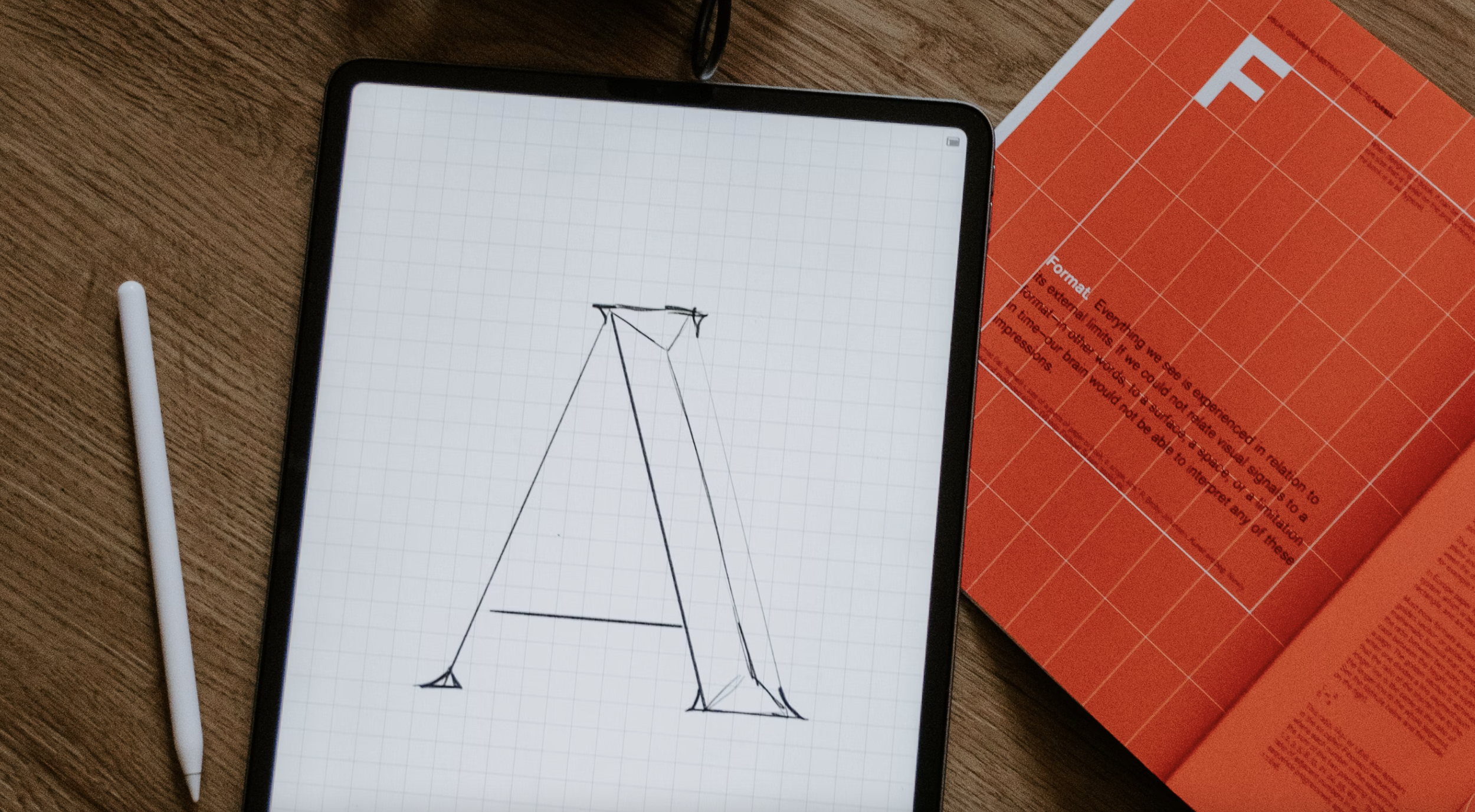 Tablet displaying letter 'A' design next to white stylus and red booklet on wooden table.