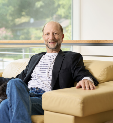 Man sitting on a beige sofa wearing a black jacket, white striped shirt, and blue jeans in a modern living room with large windows.