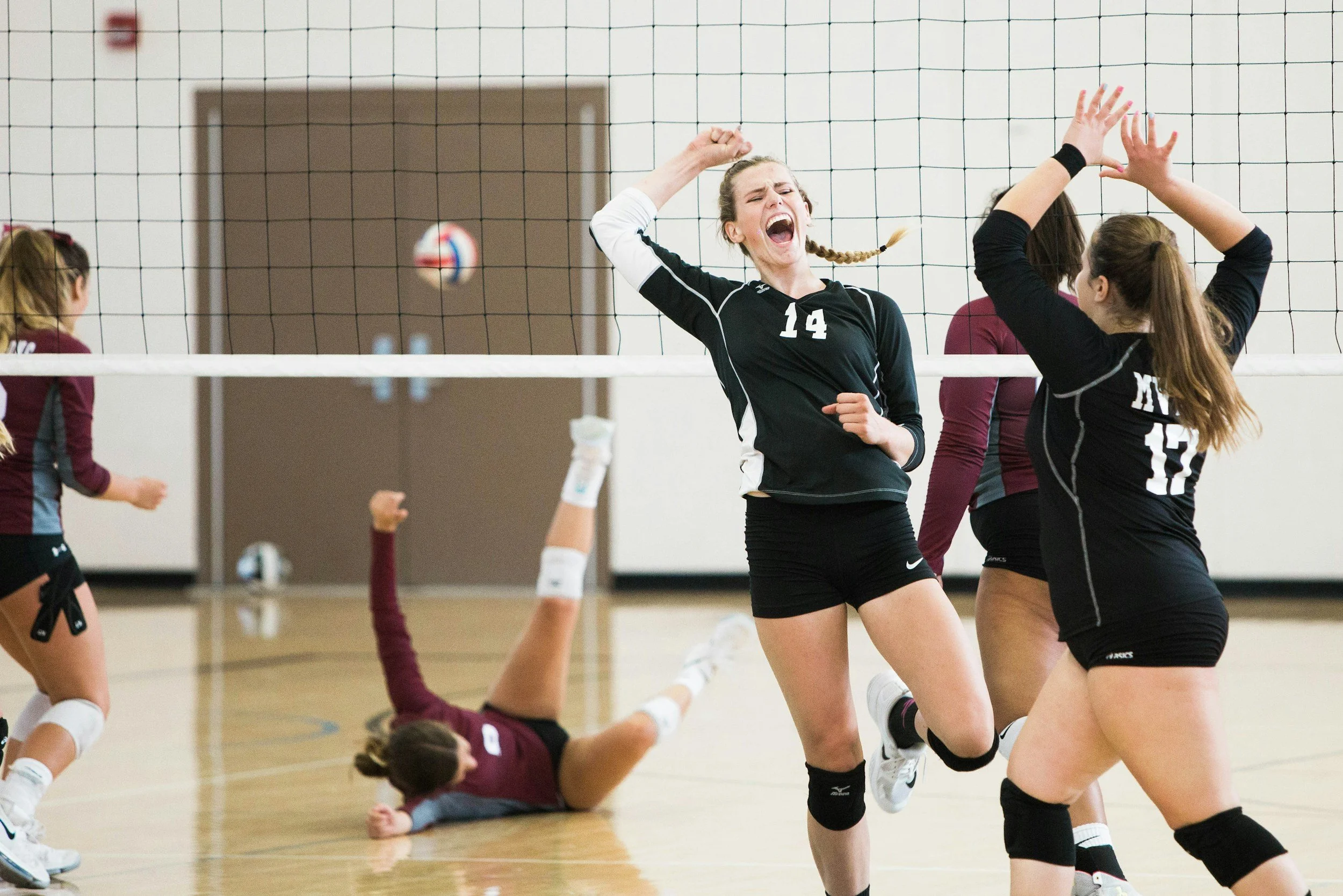A volleyball game with players celebrating a point, one player on the ground and others jumping near the net.