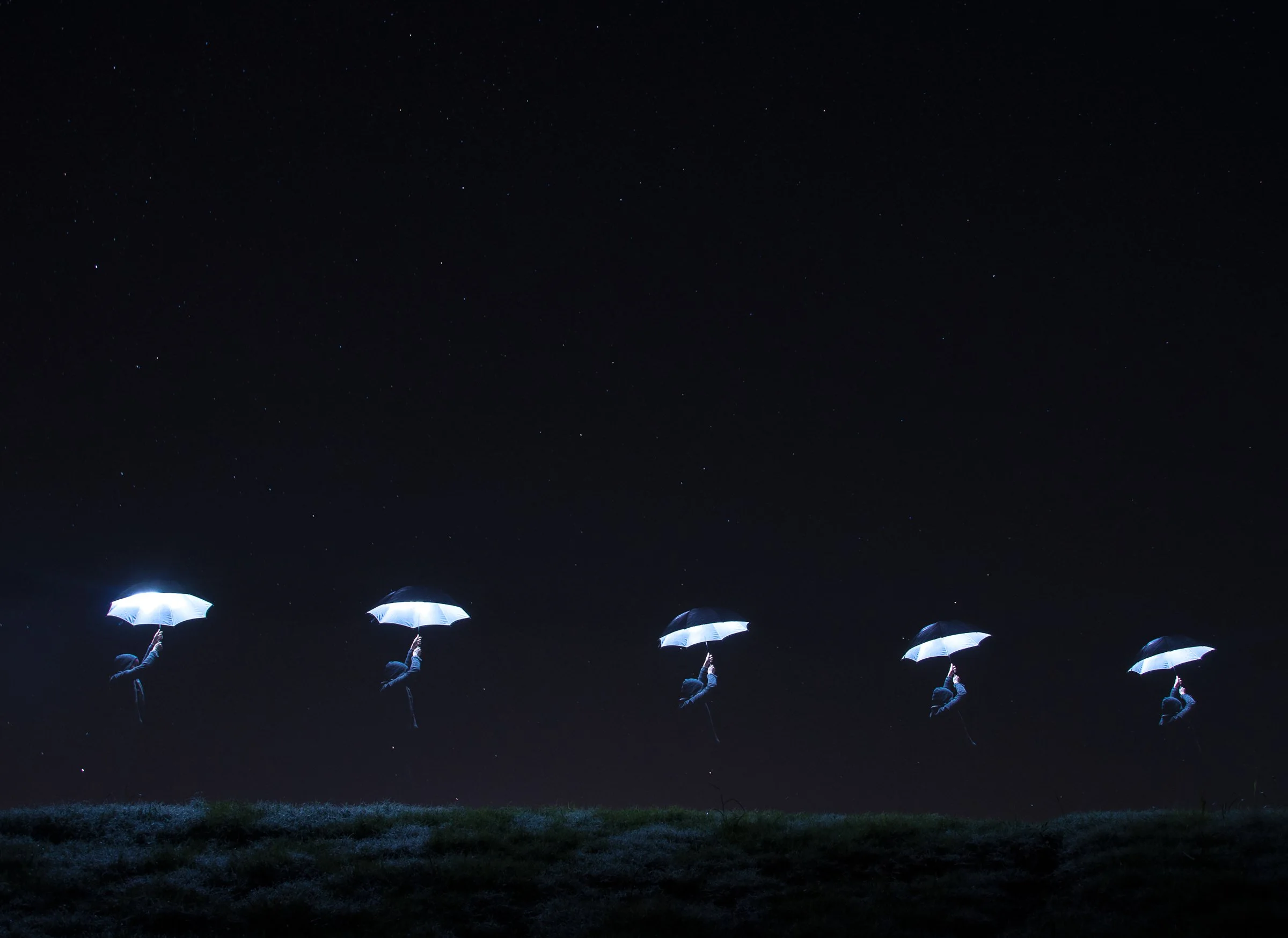 Multiple figures holding illuminated umbrellas appear to be floating in the night sky against a starry backdrop, creating an artistic and surreal scene.