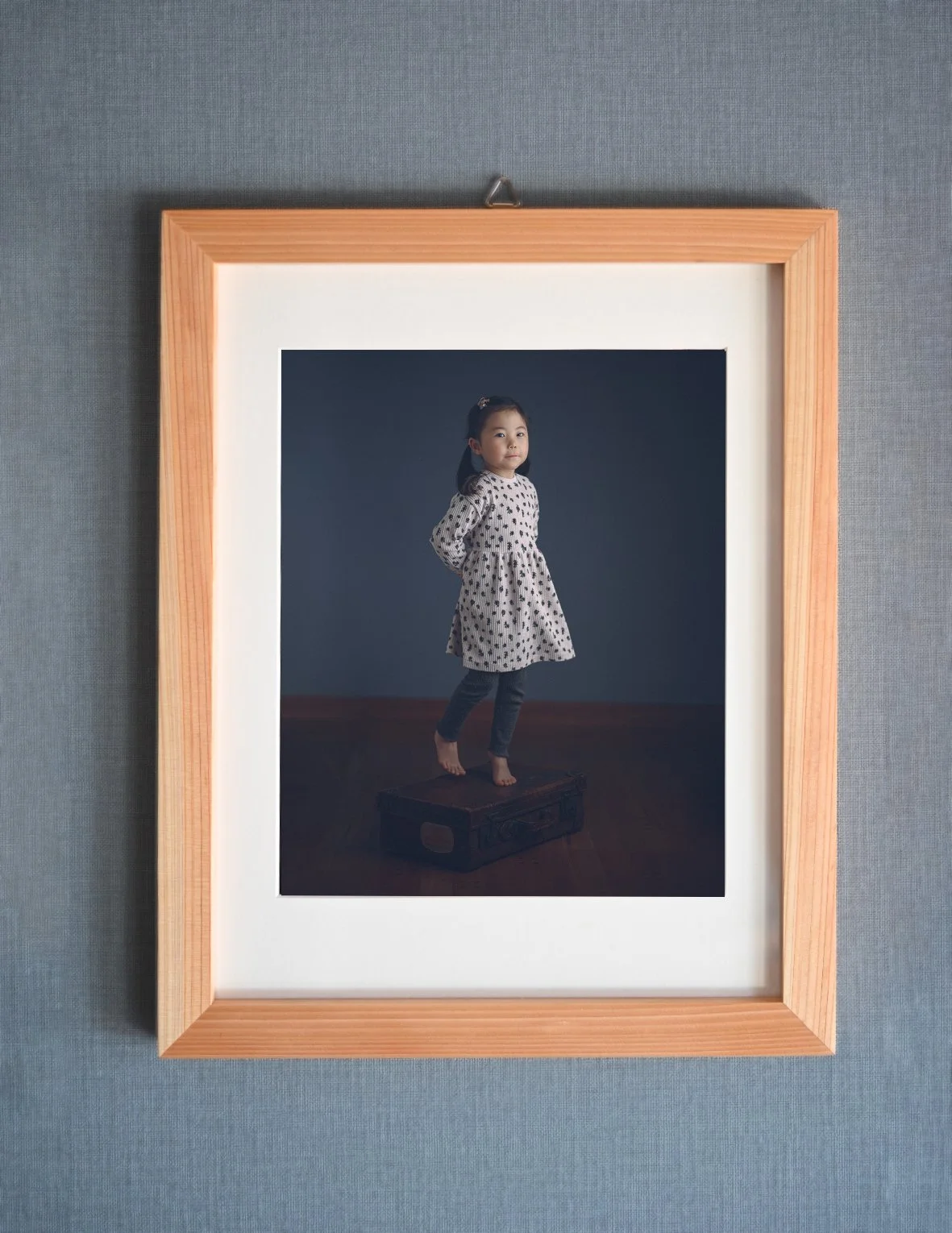 Photograph of a young girl standing on a vintage suitcase, dressed in a polka dot dress and jeans, posing against a dark background, framed and hanging on a wall.