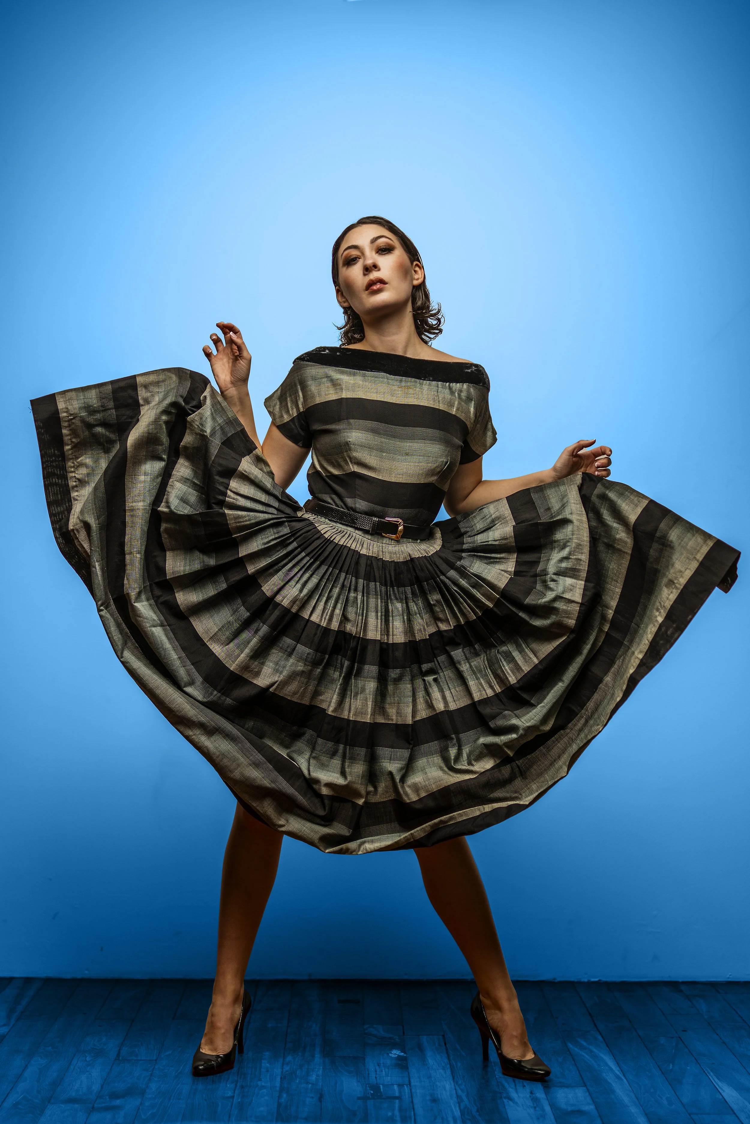 A woman in a plaid dress with a pleated skirt, wearing high heels, stands with her legs apart against a blue background, holding the sides of her dress.