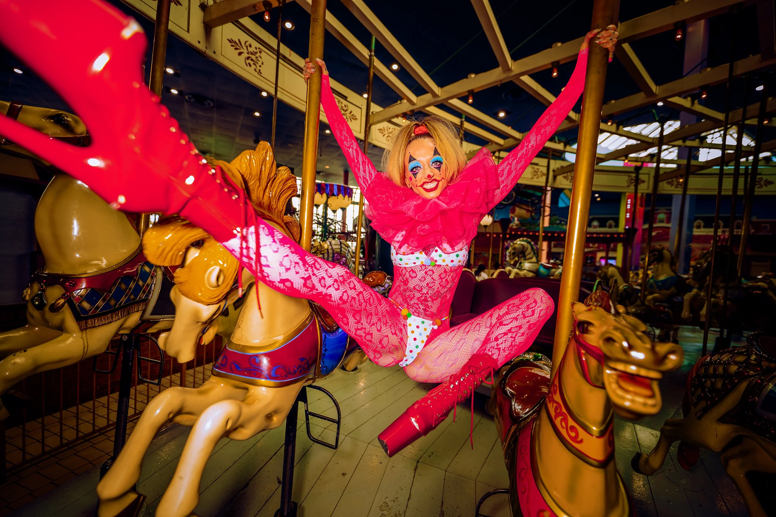 A person dressed as a clown with exaggerated makeup and a pink outfit, sitting on a carousel horse at an amusement park.