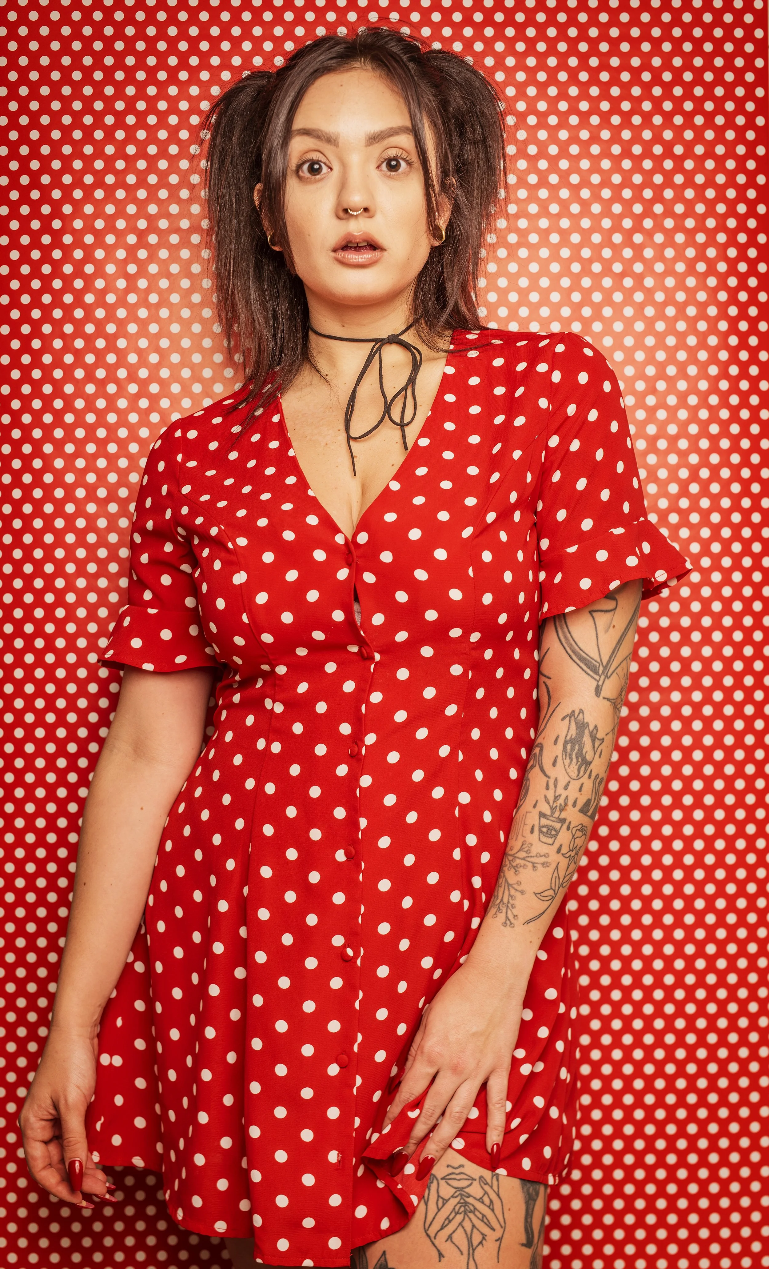 A woman in a red dress with white polka dots stands in front of a red background with white polka dots. She has dark hair styled in pigtails, tattoos on her arms, and is wearing a septum ring and a black choker. She has a surprised or startled expres
