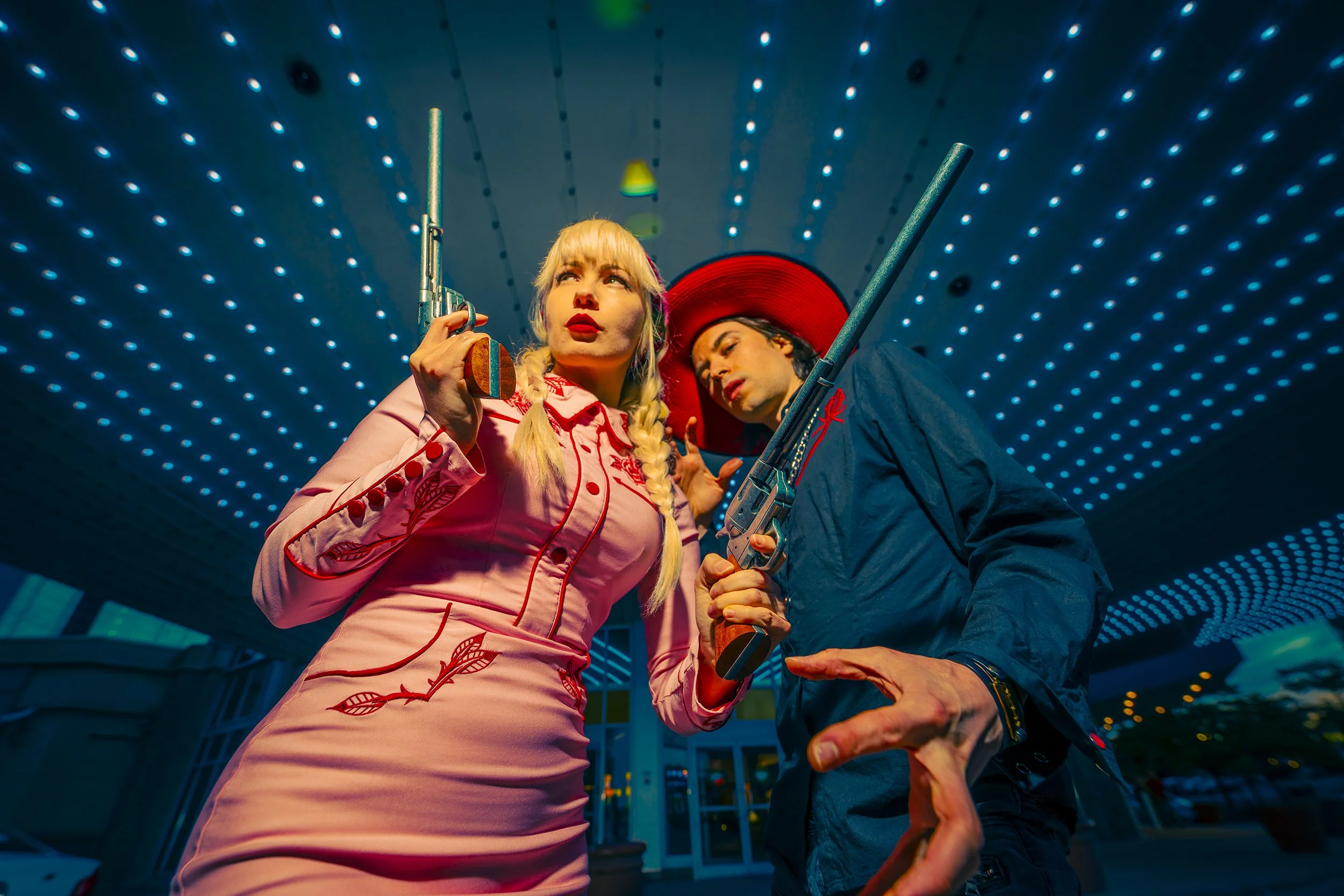 Two people are posing underneath a ceiling with numerous small blue lights. The woman on the left is wearing a pink dress with leaf patterns, has blonde hair styled in braids, and is holding a gun prop. The man on the right is wearing a dark jacket w