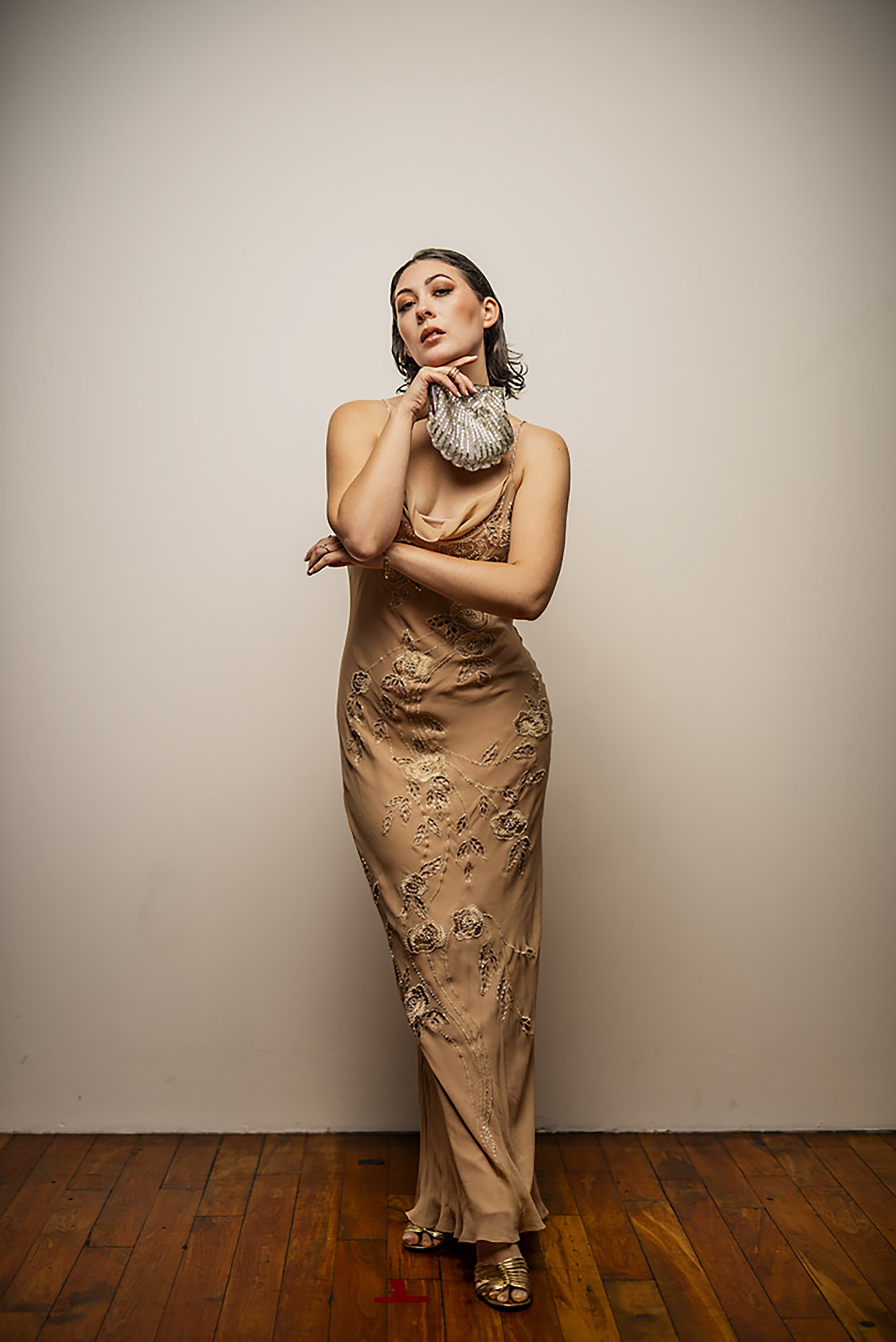 A woman with dark, shoulder-length hair and makeup, dressed in a beige, embroidered, silk gown, holding a silver clutch, standing indoors on a wooden floor against a plain light-colored wall.