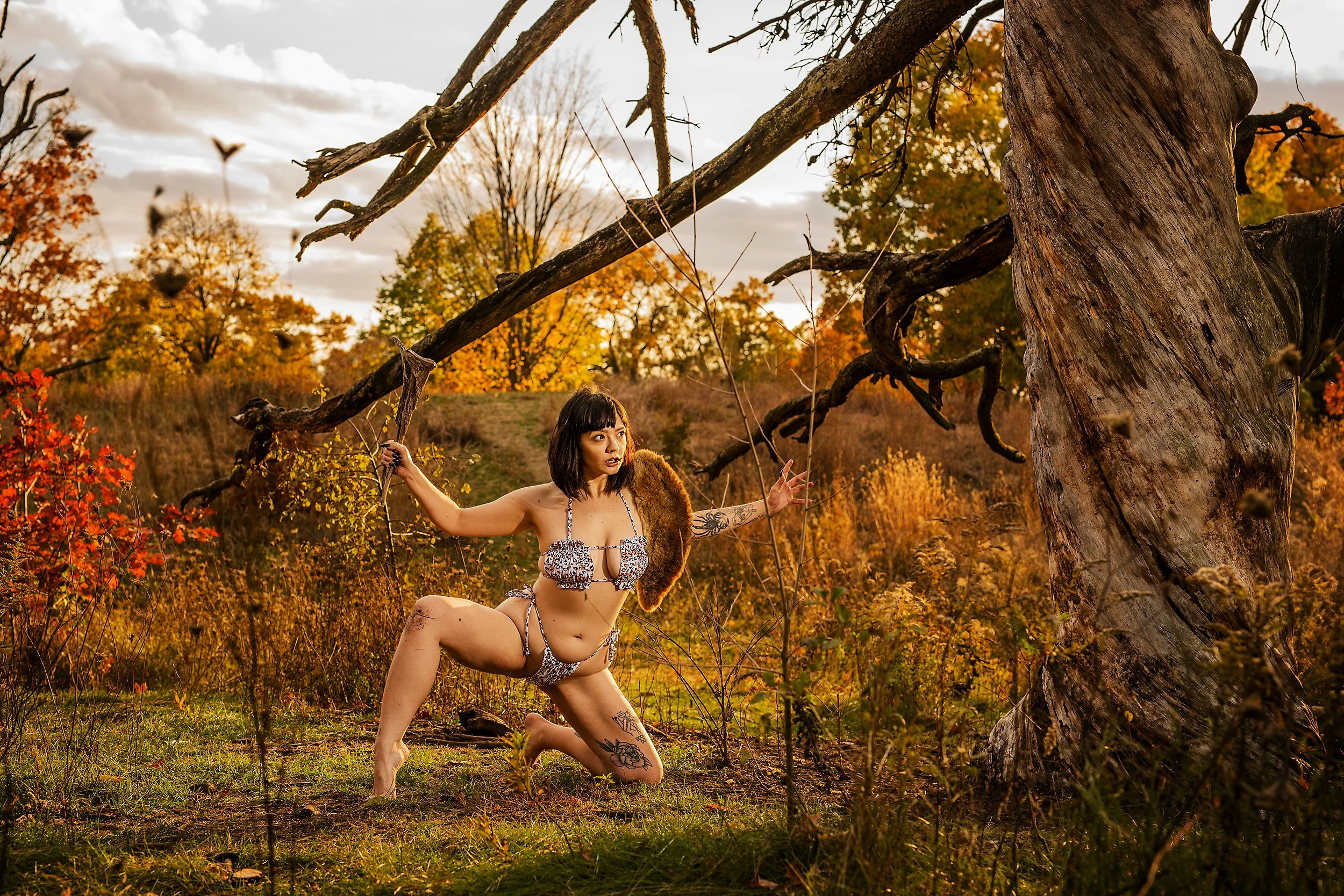 A woman in leopard print bikini and tribal accessories in a forest with autumn foliage, kneeling on one knee, reaching out and posing near a large, twisted tree.