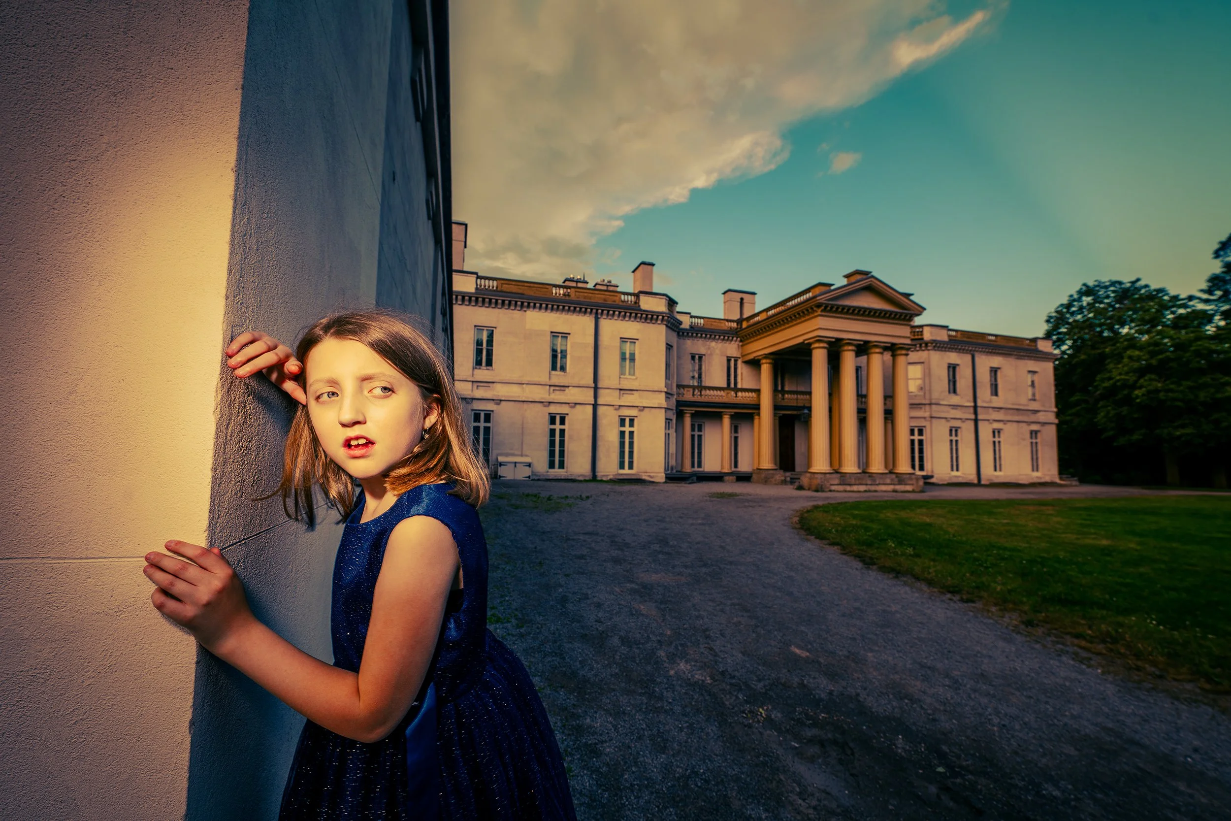Young girl in a blue dress with long hair leaning against a textured wall at sunset, with a historic building with columns and a portico in the background.