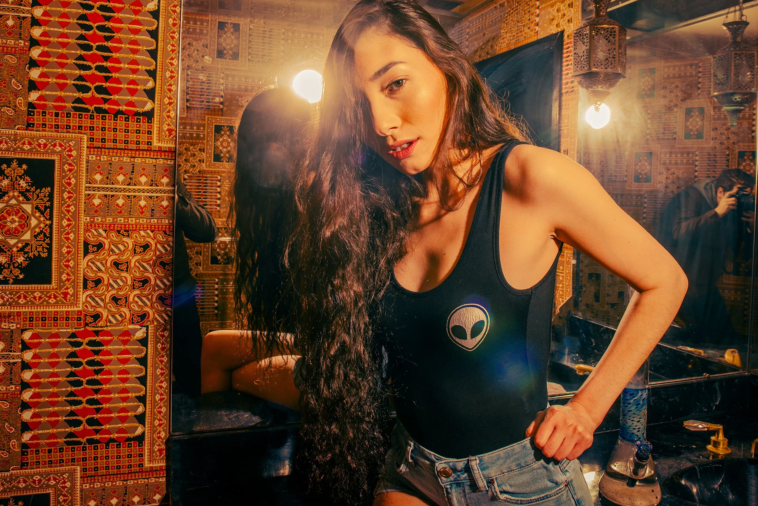 A woman with long dark hair smiling slightly, wearing a black tank top with a white alien face logo, standing in front of a mirror with ornate, colorful rugs hanging on the wall behind her.