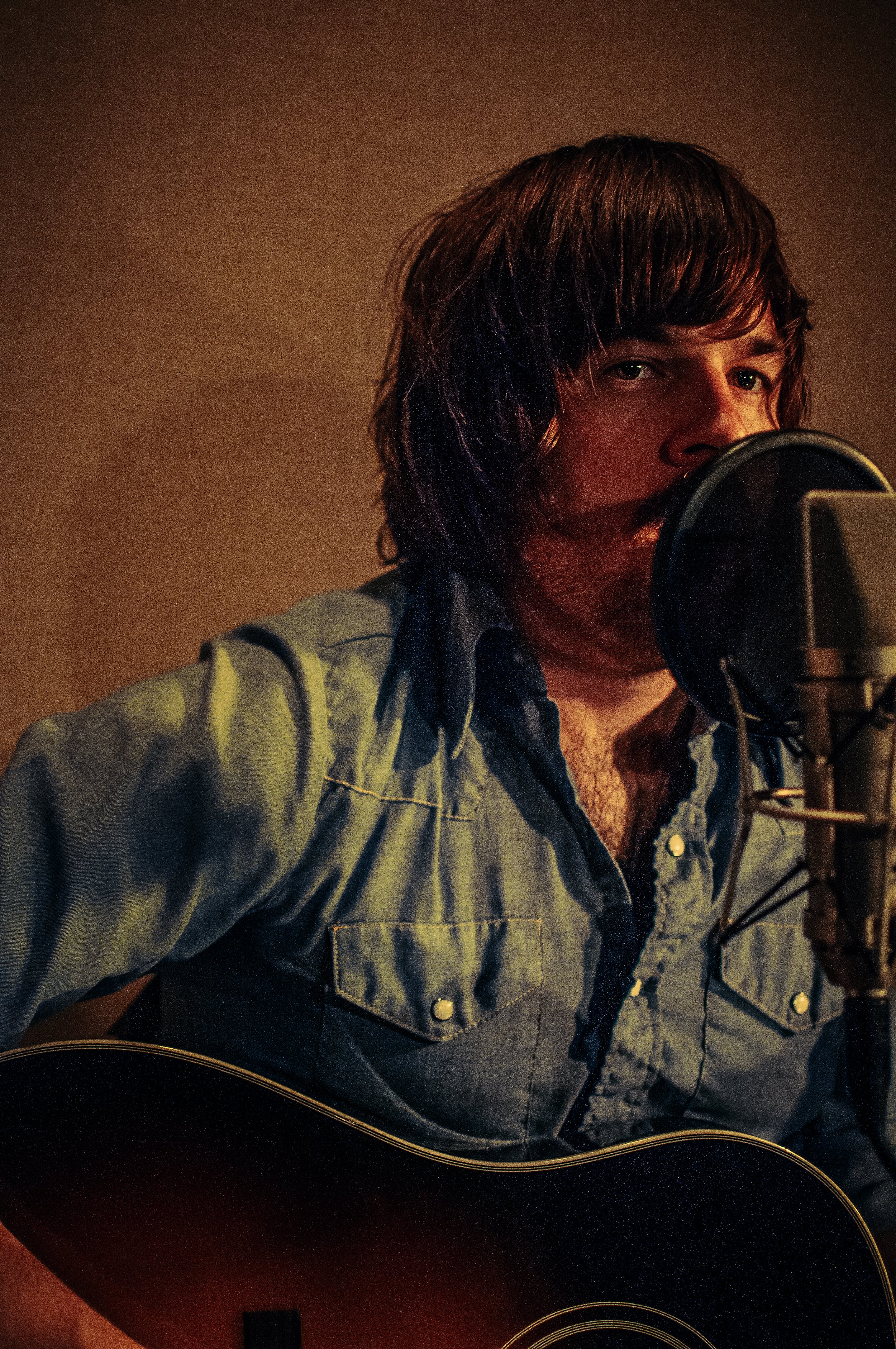 A man with brown hair and a beard is singing into a microphone while playing an acoustic guitar. He is wearing a denim shirt and looking towards the camera.