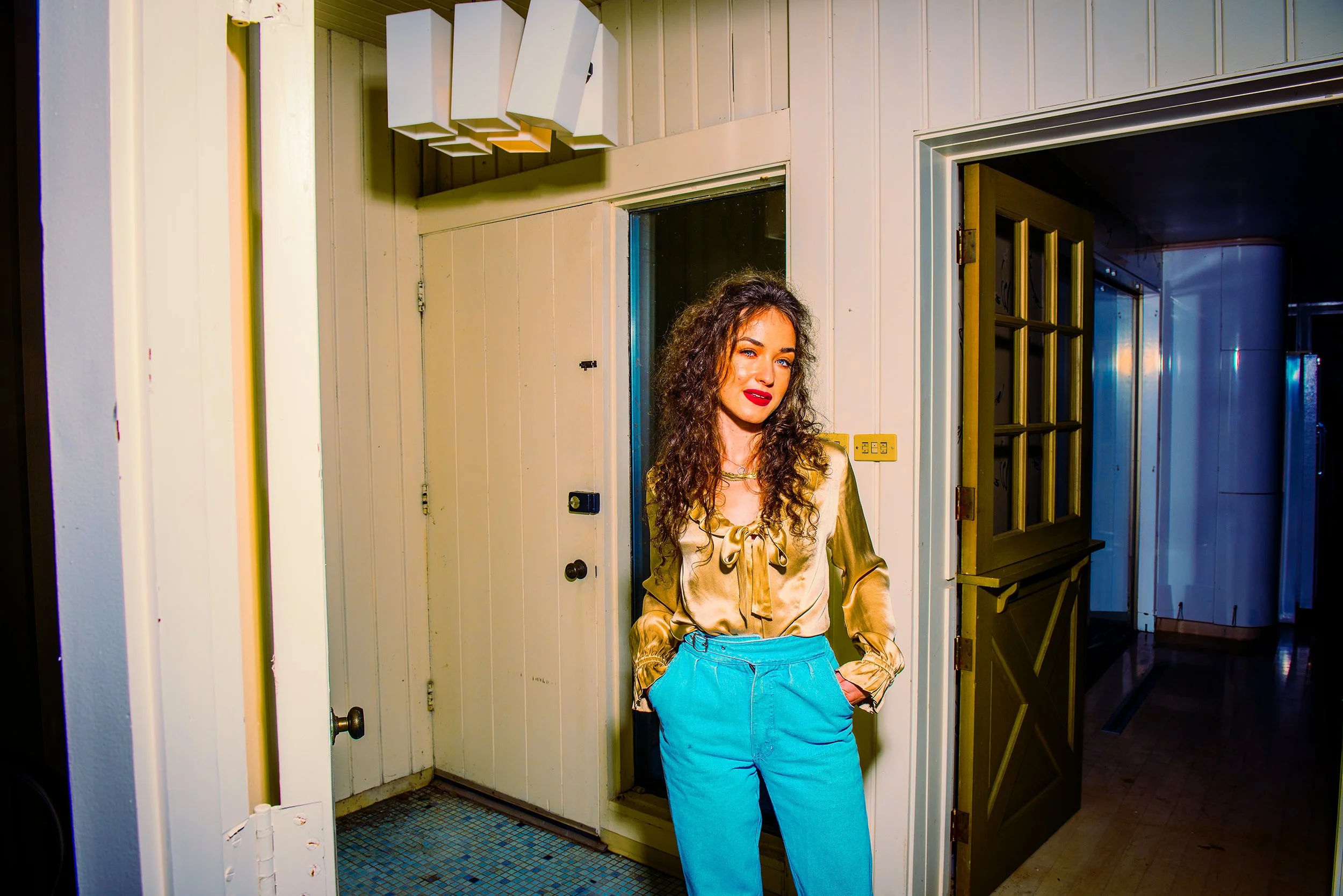 A young woman with curly hair wearing a satin gold blouse and blue jeans, standing in a doorway inside a building with white paneled walls and tiled floor.